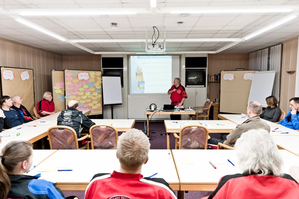 Ein Lehrer in einer roten Uniform spricht vor einer Gruppe von Erwachsenen in einem Seminarraum, der mit Moderationswänden und einem Whiteboard ausgestattet ist.