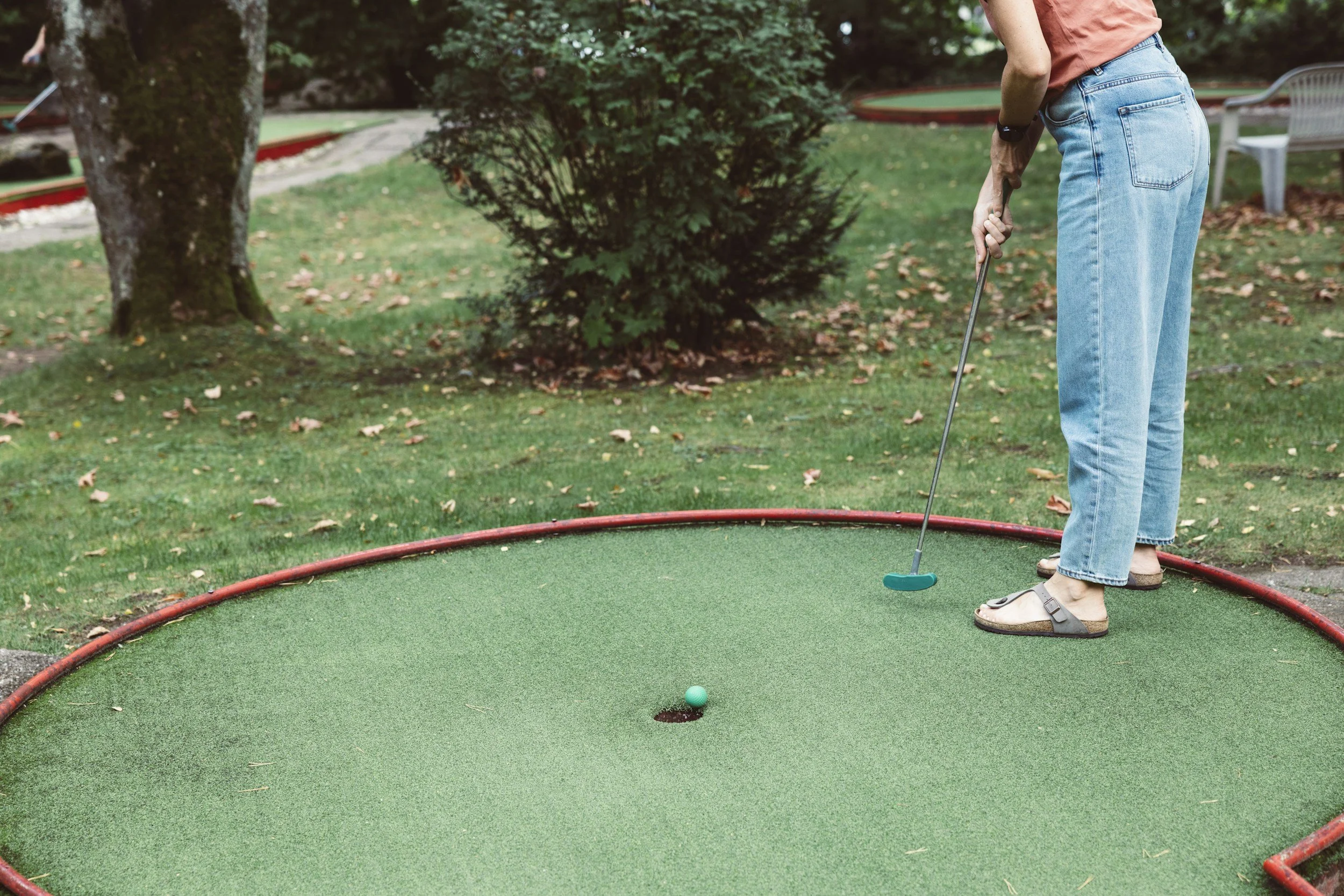 Person, der Mini-Golf spielt, trägt pinkfarbenes T-Shirt und blaue Jeans, spielt auf einem grünen Mini-Golf-Feld im Freien, umgeben von Bäumen und Bänken.