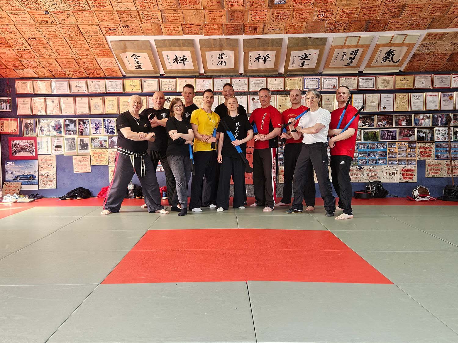 Gruppe von Menschen in einem Kampfsportstudio, die für ein Gruppenfoto posieren, einige mit Kampfsportwaffen, im Hintergrund Wände mit Zertifikaten, Fotos und Kampfsportzeichen.