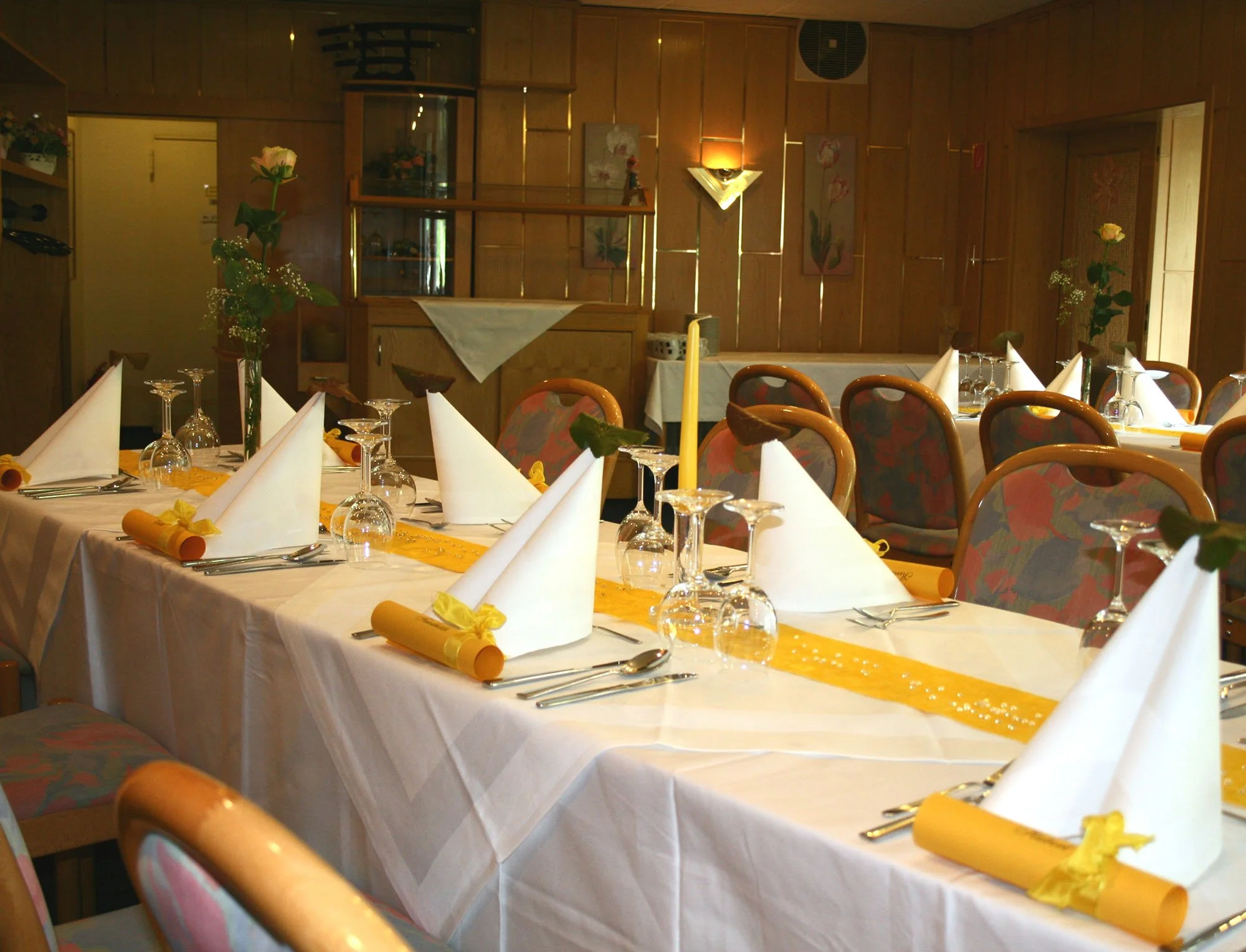 Ein festlich gedeckter Esstisch mit weißen Tafeldecken, weißen Stoffservietten, gelben Papierrequisiten und Kristallweingläser in einem Restaurant mit Holzinterieur und Blumenvasen.
