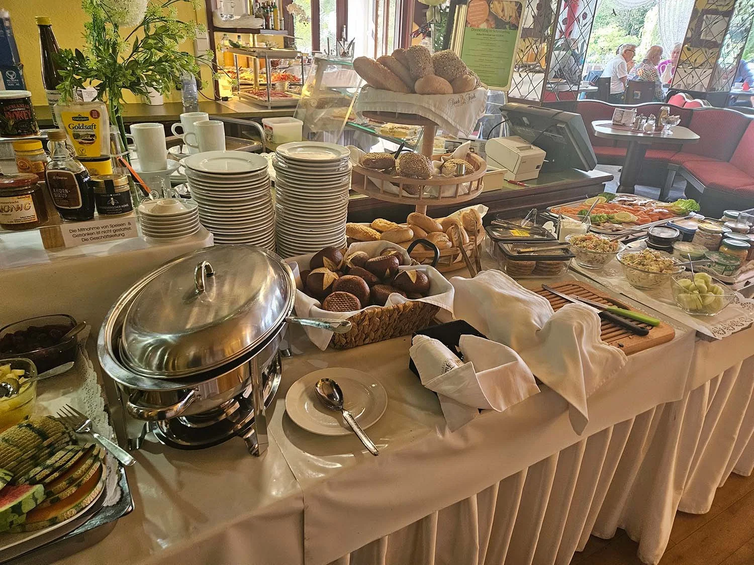 Frühstücksbuffet mit Broten, Croissants, Aufschnitt, Salaten, Joghurt und diversen Getränken in einem Café.