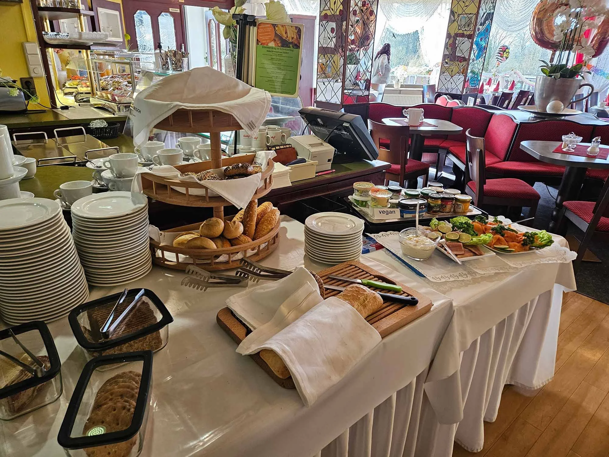 Ein Frühstücksbuffet mit Tellern, Gebäck, Joghurt, Salat und weiteren Speisen im Themenhotel Bad Münder mit großen Fenstern und roten Sitzmöbeln im Hintergrund.