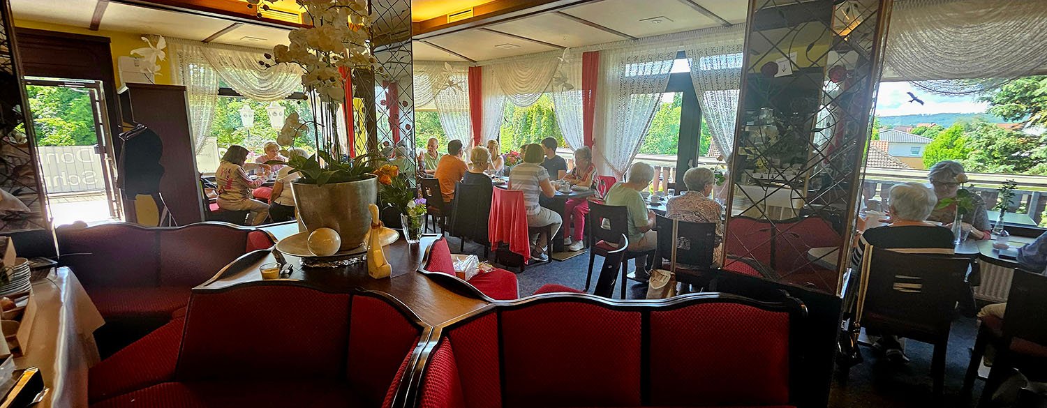 Ein belebtes Restaurant mit mehreren Tischen, an denen ältere Menschen essen. Es gibt große Fenster mit Blick nach draußen auf grüne Bäume und Häuser. Die Einrichtung ist dekorativ mit Blumen und Möbeln in Rot und Dunkelbraun.