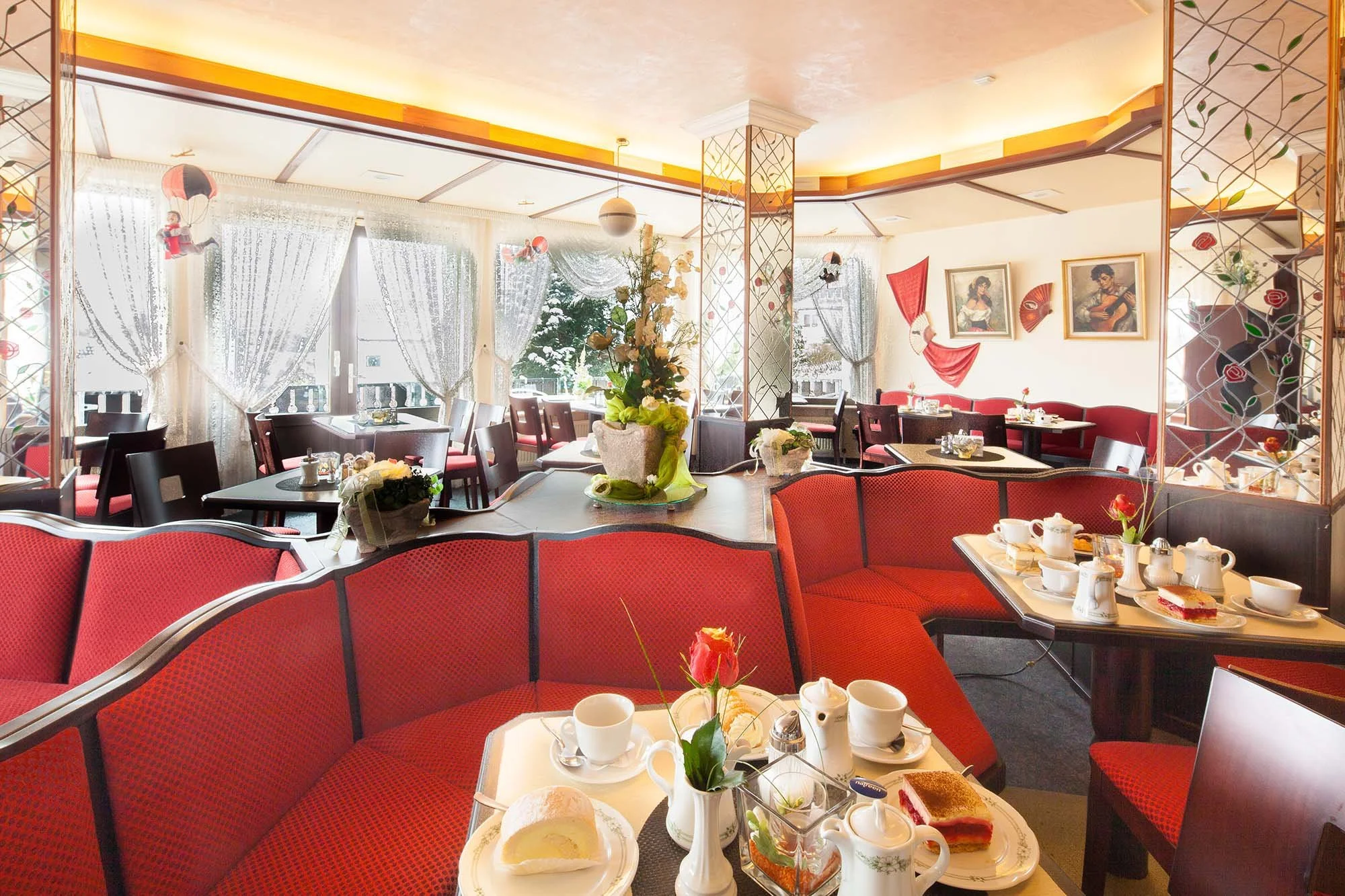 Ein elegantes Restaurant mit roten Stühlen, Gesellschaftstischen, weißem Geschirr, Tee- und Kaffeetassen, Blumenarrangements, geschnitzten Dekorationen, gemalten Porträts und einer großen Fensterfront