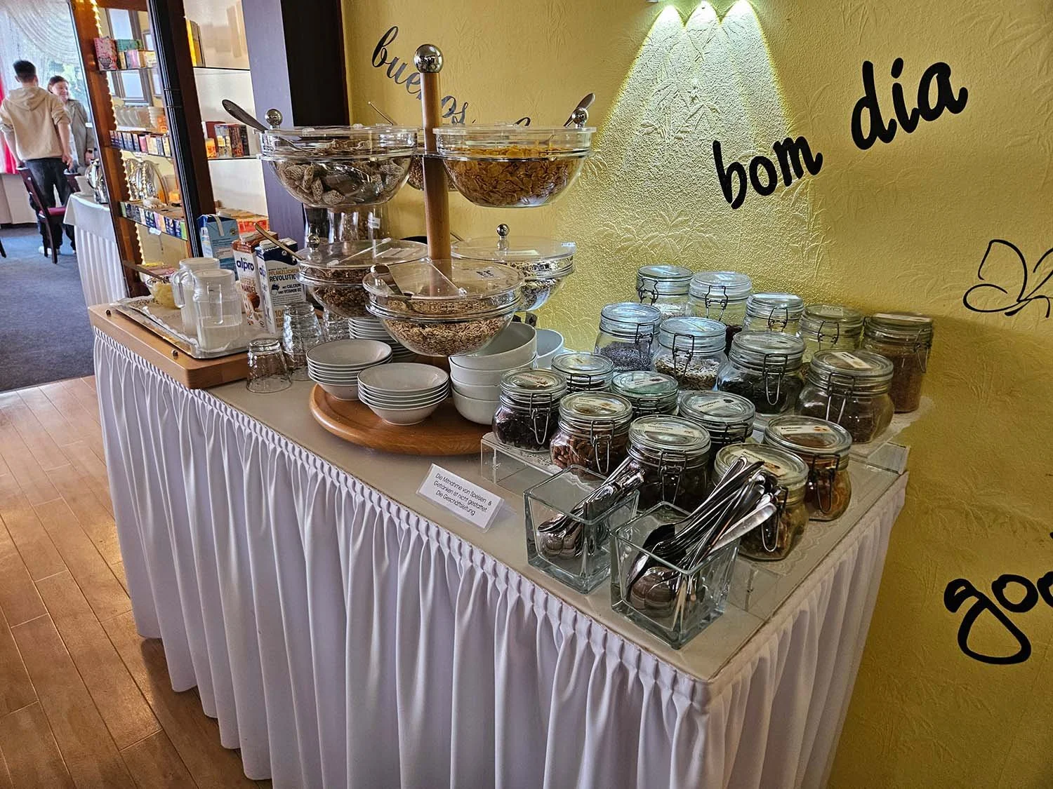 Frühstücksbuffet mit verschiedenen Müslis, Joghurt und Toppings in Gläsern, Tassen, Schüsseln und Besteck auf einem weißen Tisch mit Stoffverkleidung, gelbe Wand im Hintergrund mit deutschem Text.