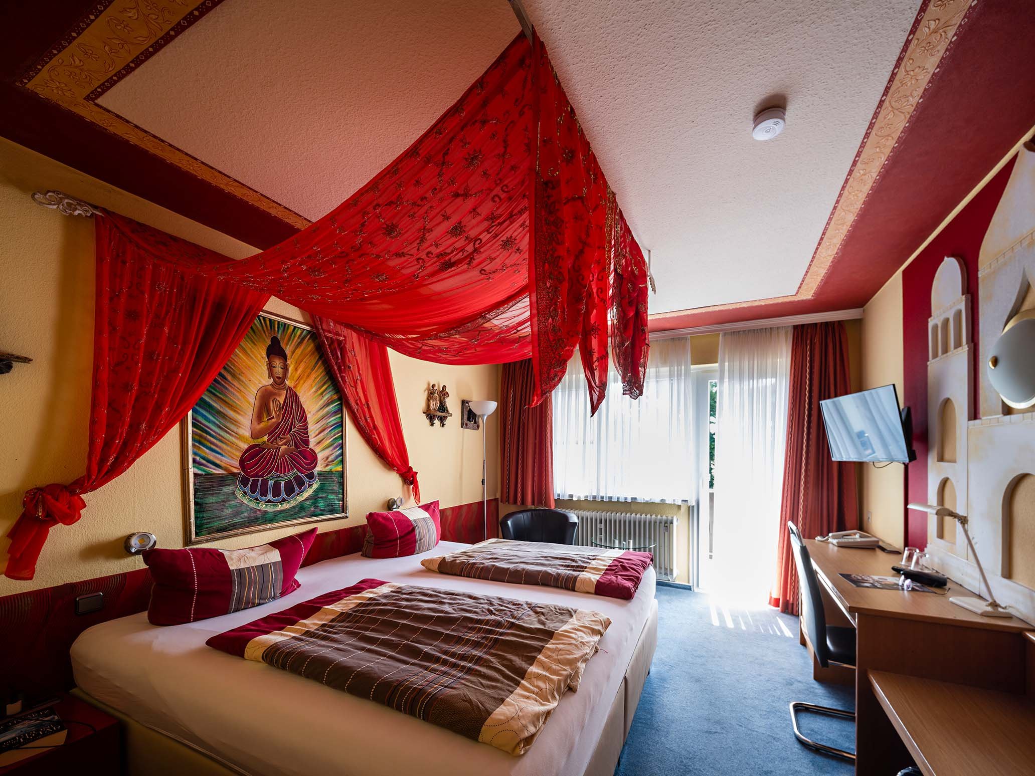Ein Hotelzimmer mit einem Bett, roten Vorhängen und Wanddekorationen, darunter ein Bild von Buddha, sowie ein Schreibtisch und ein Fernseher.
