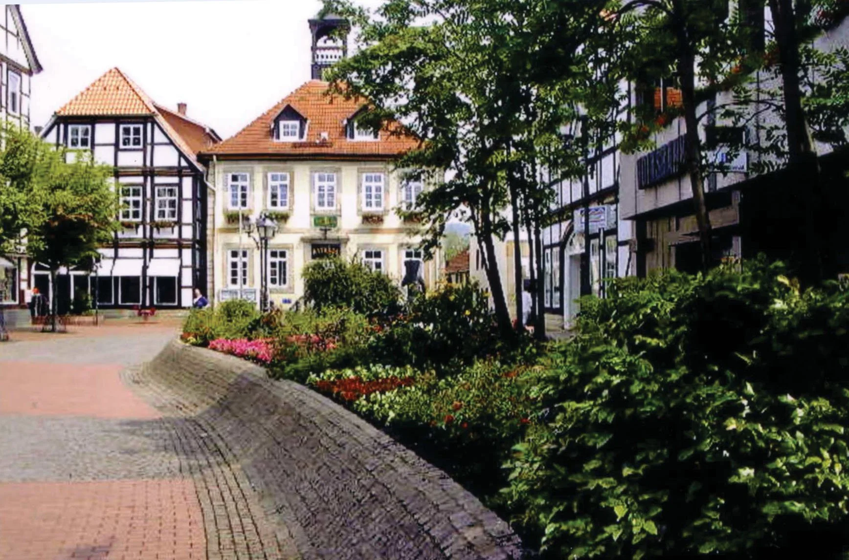 Ein malerischer Stadtplatz mit Blumenbeeten, historischen Fachwerkhäusern und Bäumen, bei sonnigem Wetter.