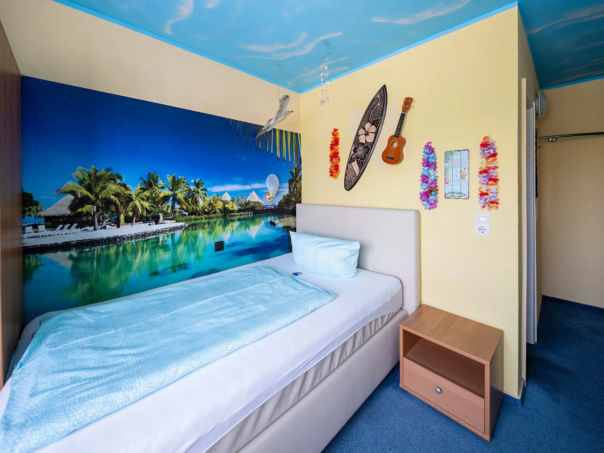 Schlafzimmer mit Wanddeko im tropischen Stil, Meer und Palmen, ein Bett mit weißer Bettwäsche, ein Nachttisch aus Holz, bunte Blumenketten an der Wand, ein kleines Klavier, eine Surfbrett-Dekoration, und eine Decke mit Himmel-Design an der Decke.
