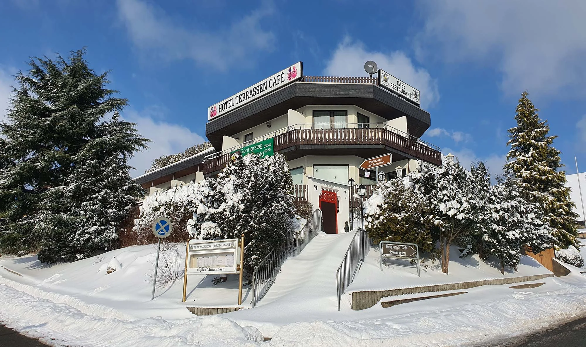 Mehrstöckiges Hotel Cafe mit Winterlandschaft im Vordergrund, Schneebedeckte Bäume und Wege, Beschilderung für Restaurant und Hotelzugang, blauer Himmel mit wenigen Wolken.