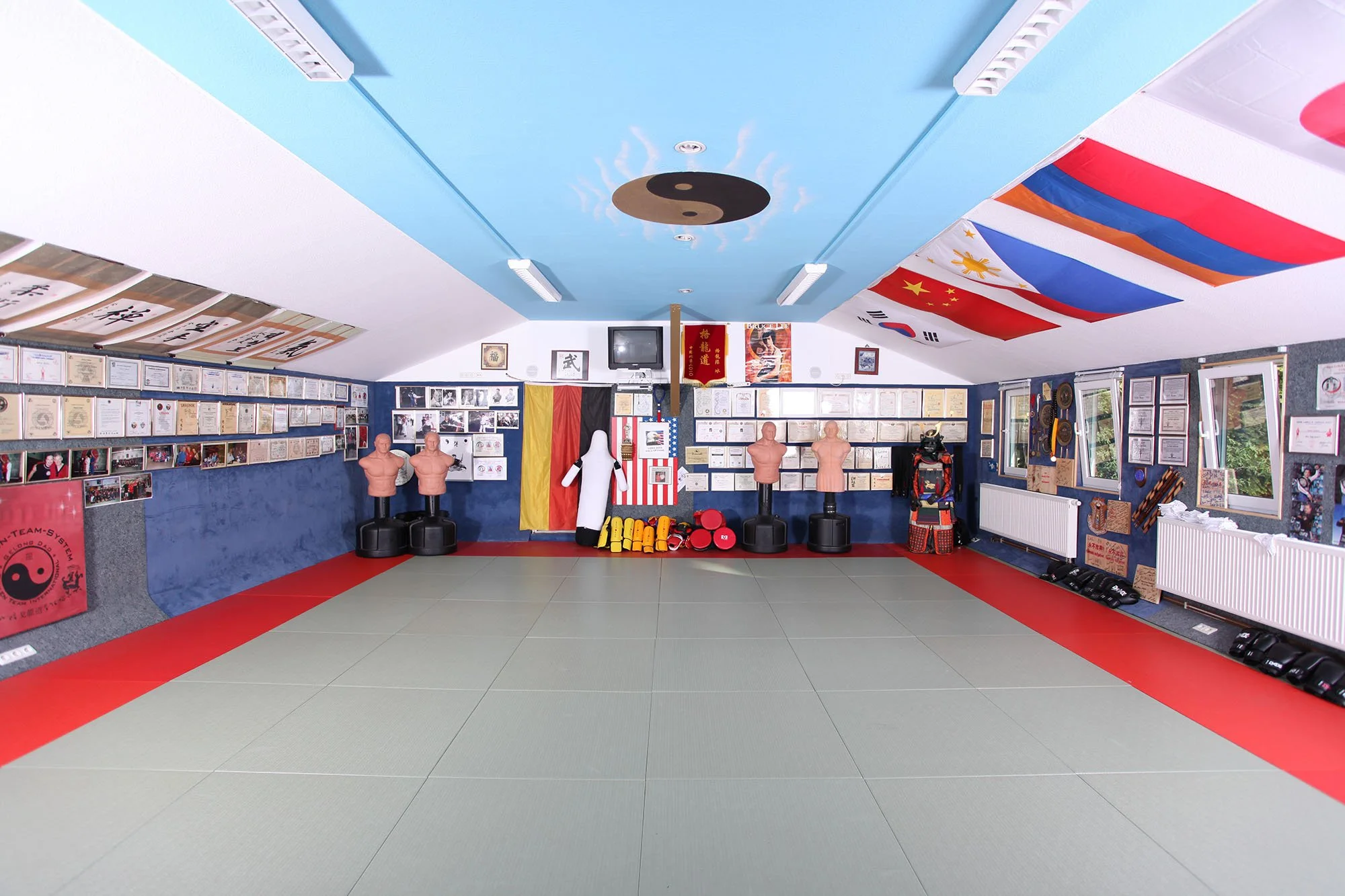 Kampfkunstraum mit Kampfsportausrüstung, Mannequins, Flaggen, Fotos, und Flaggen an der Wand, im Mittelpunkt ein Übungsmatte, an den Seiten Fenster