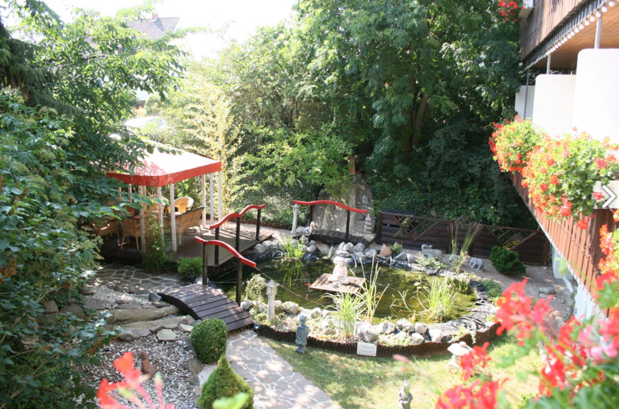 Garten mit Teich, Steinen, Bänken, Blumentöpfen und einer überdachten Terrasse auf der rechten Seite.