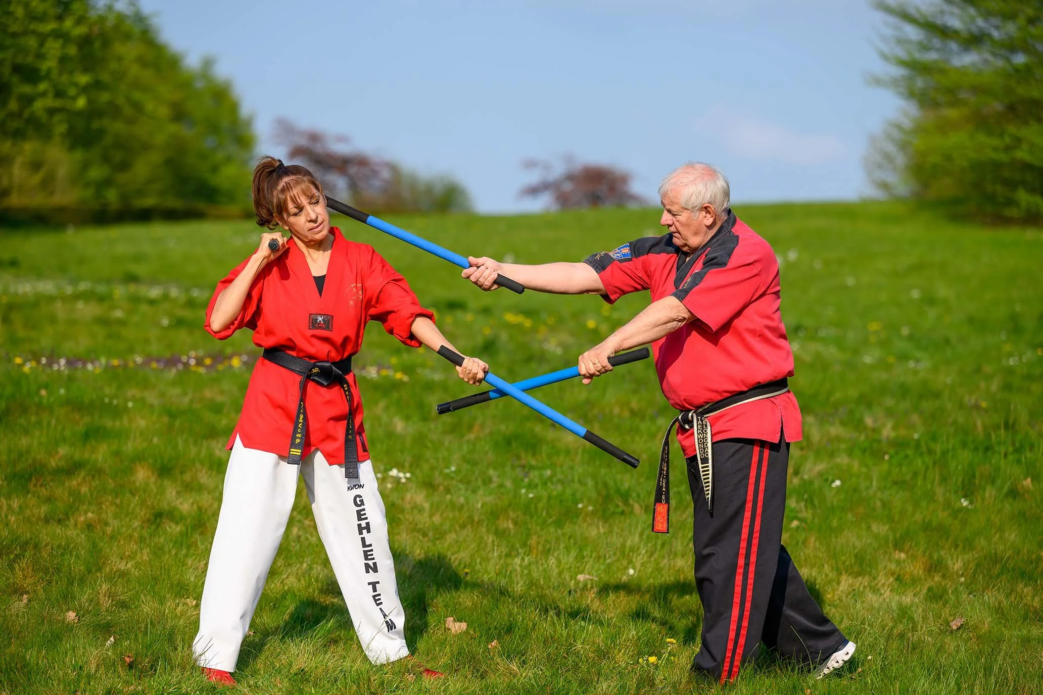 Zwei Karate-Kämpfer, eine Frau und ein Mann, üben im Freien. Beide tragen rote Karate-Kleidung, der Mann schlägt mit einem Stock. Sie stehen auf grünem Gras, im Hintergrund Bäume und blauer Himmel.