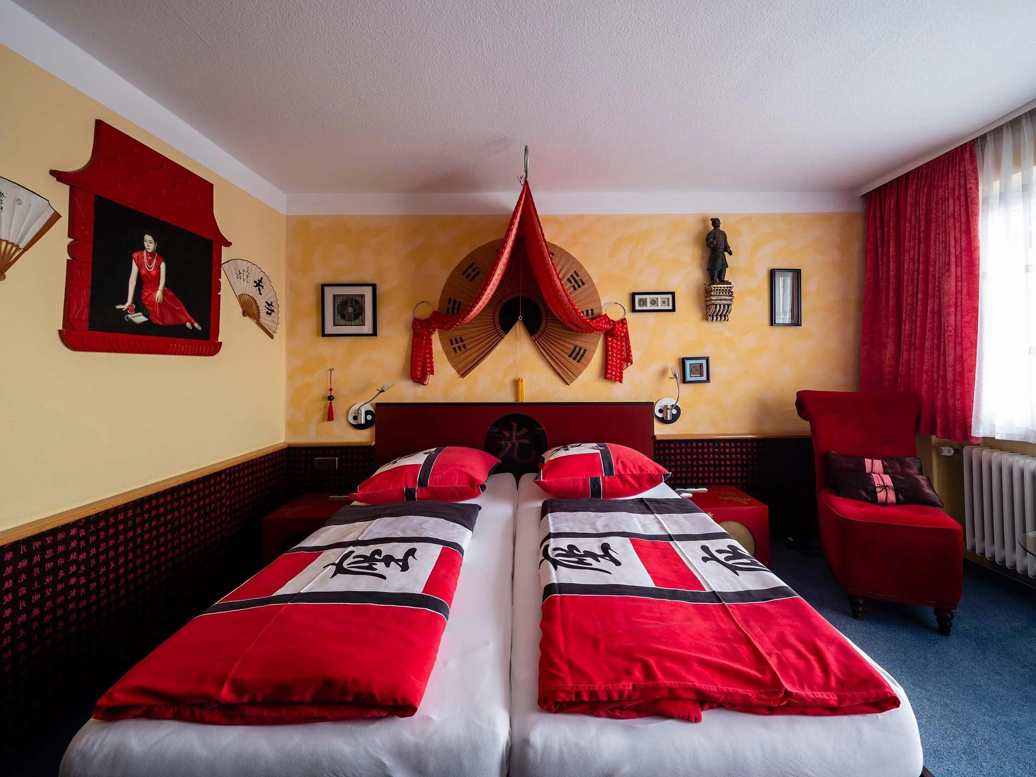 Ein Schlafzimmer mit chinesischer Dekoration, inklusive Wandbildern, Fächer und roten Vorhängen, zwei Betten mit schwarz-weißen Bettdecken und roten Kissen.