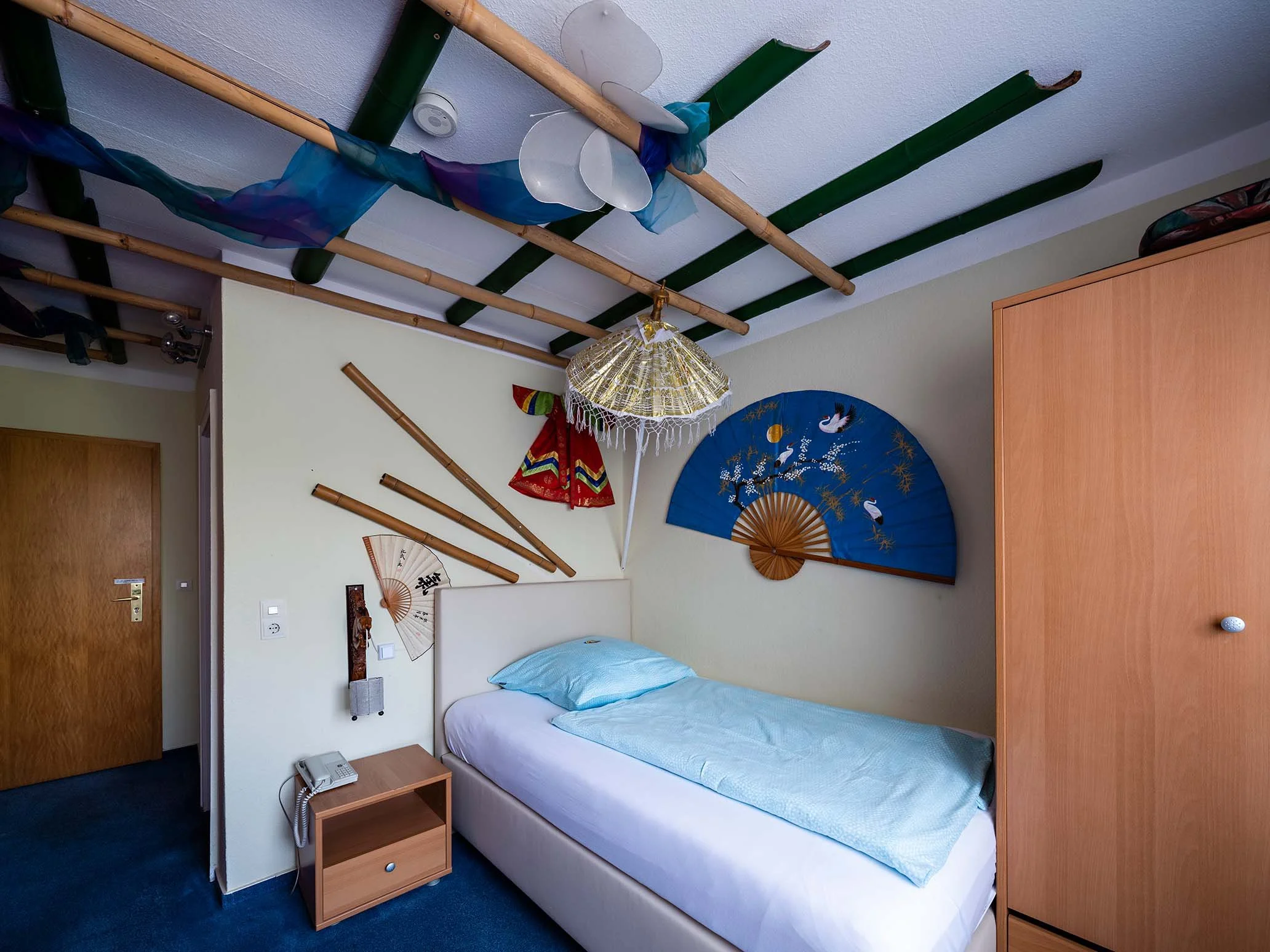 Ein Gästezimmer mit einem Einzelbett, einem Nachttisch mit Telefon, an der Wand hängen japanische Fächer, eine Deckenlampe und ein Hausflur vor der Tür.
