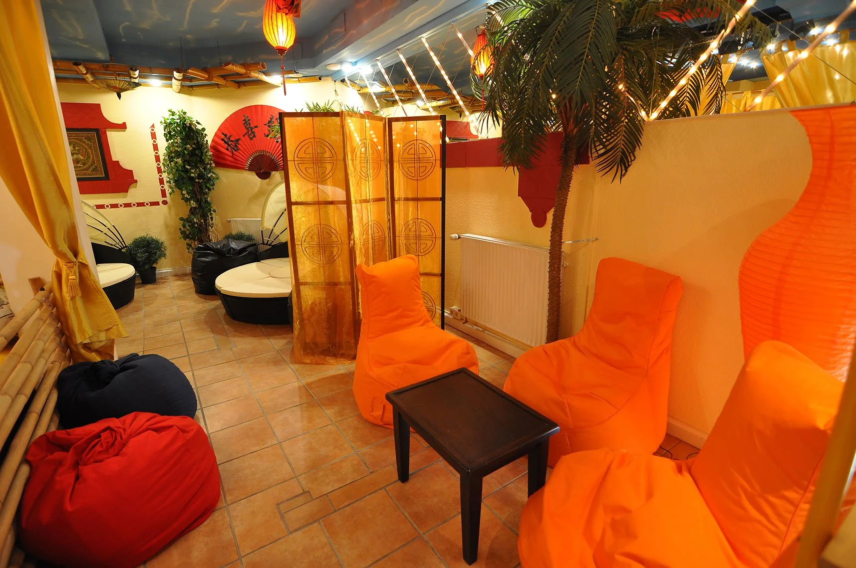 Ein gemütlich eingerichteter Raum mit orangefarbenen Sitzsäcken, einer Raumtrennwand mit chinesischen Mustern, Pflanzen und Dekoration mit asiatischem Stil, bunte Beleuchtung und eine Wandlampe.