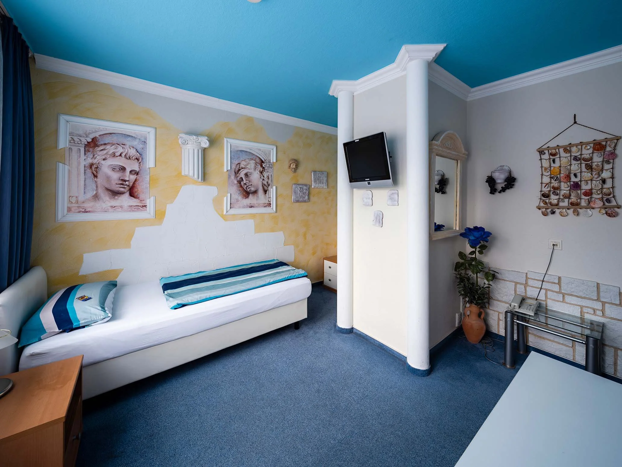 Ein Hotelzimmer mit einem weißen Bett, künstlerischen Wanddekorationen, einem Fernseher und einem kleinen Tisch, mit blauen und gelben Farbtönen und klassischen Elementen.