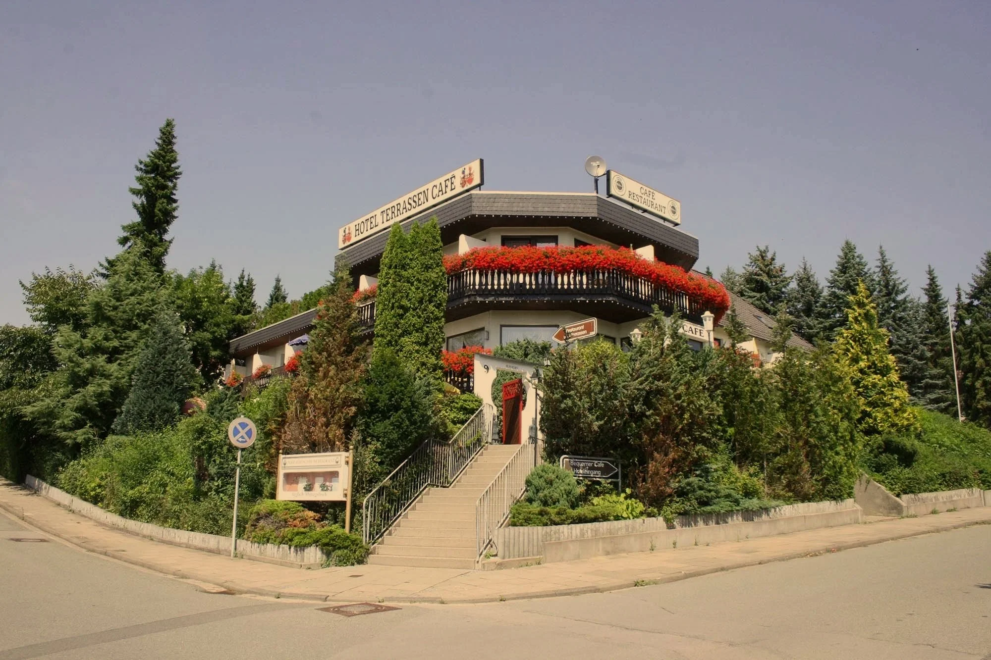 Mehrstöckiges Hotel mit Terrasse und Garten, umgeben von Bäumen und Pflanzen, mit Treppe und Straßenecke im Vordergrund.