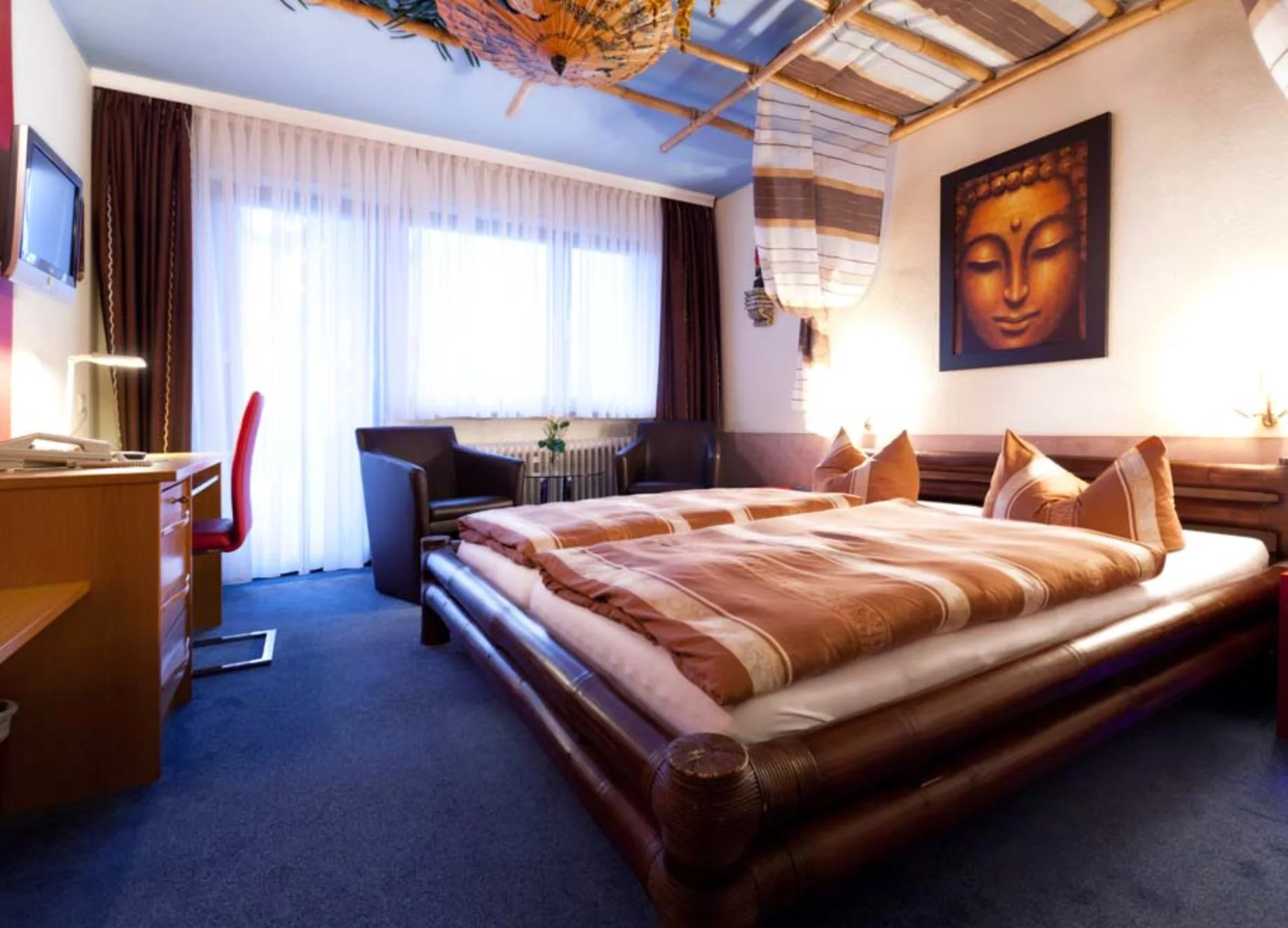 Ein Hotelzimmer mit zwei Betten, einer Sitzgruppe, einem Wandbild eines Buddha, einem Fernseher, Vorhängen vor einem großen Fenster und Dekorationen im asiatischen Stil.