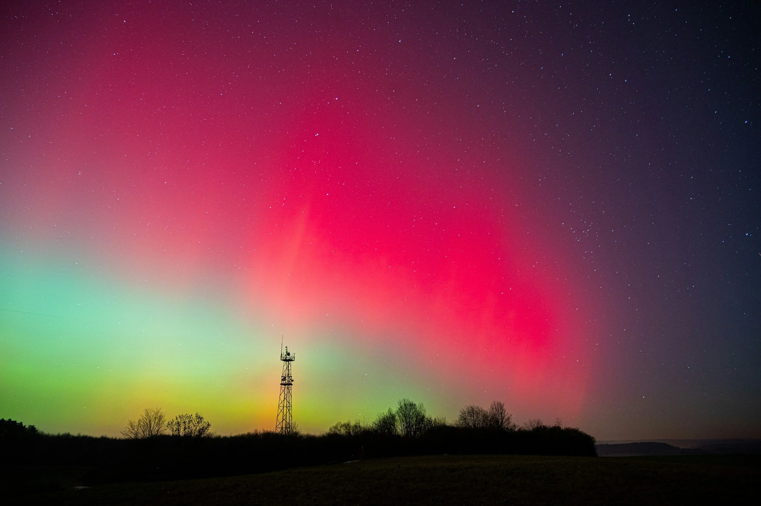 Northern Lights (Aurora Borealis) in pink, green, and purple hues over a landscape with silhouetted trees and a communications tower at night.