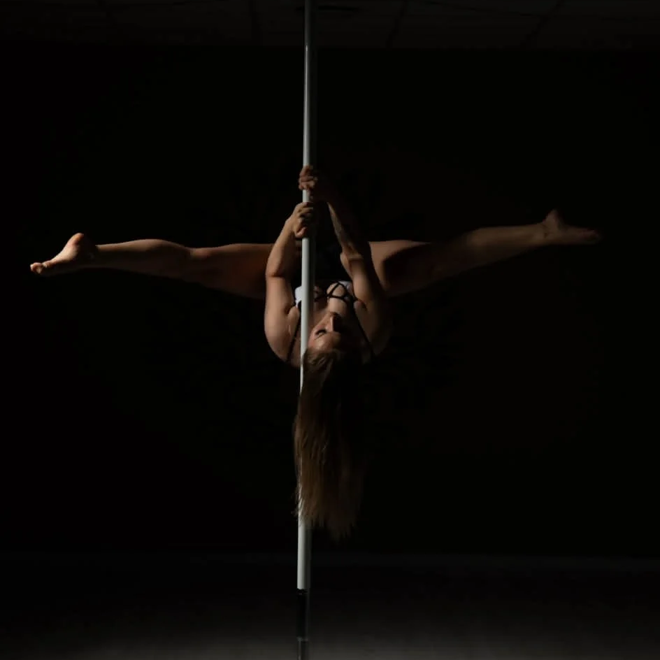 A person performing a pole dance, hanging upside down with legs spread wide, in a dark setting with minimal lighting.