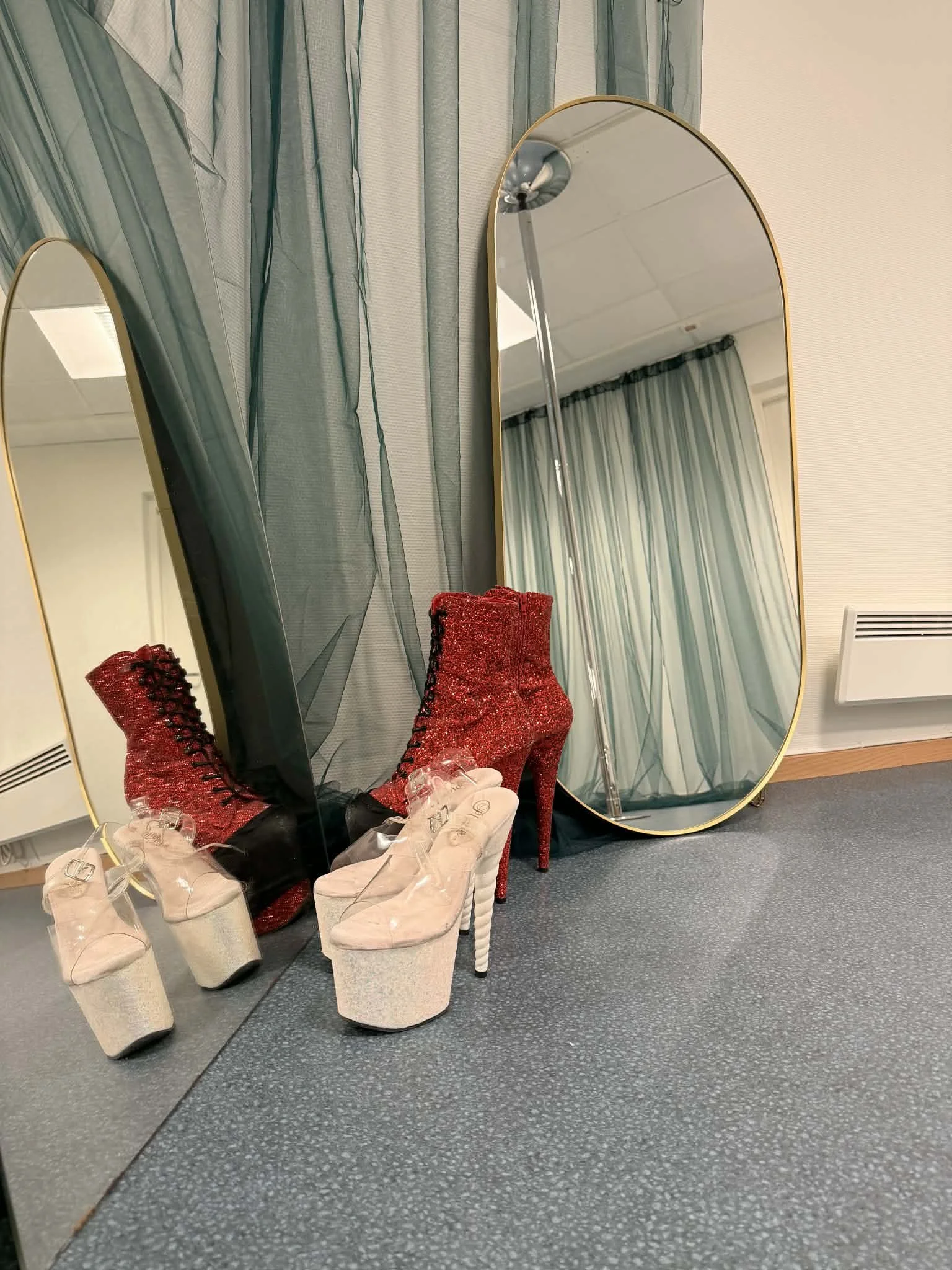 Reflection of red glitter high-heeled boots and beige platform shoes with clear heels in two standing mirrors, with blue curtains and a white wall in the background.