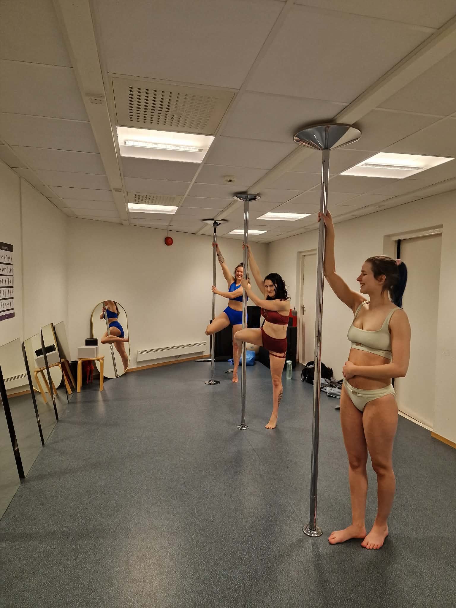 Three women in workout clothes practicing pole dancing in a room, with mirrors on the wall, some standing on the floor and one using the pole with a leg hook.