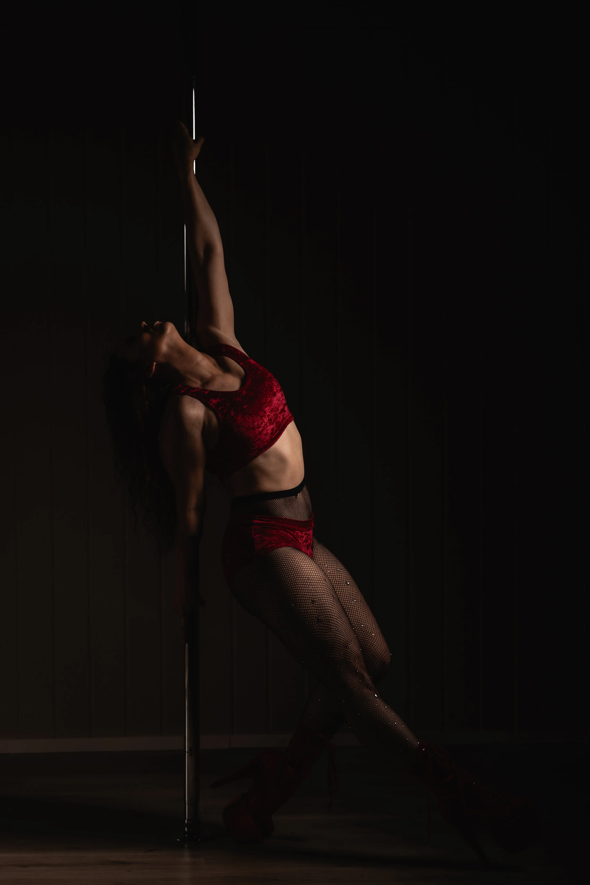 A woman in a red pole dance outfit performs a graceful pose on a vertical pole, with one leg extended upward and the other bent.