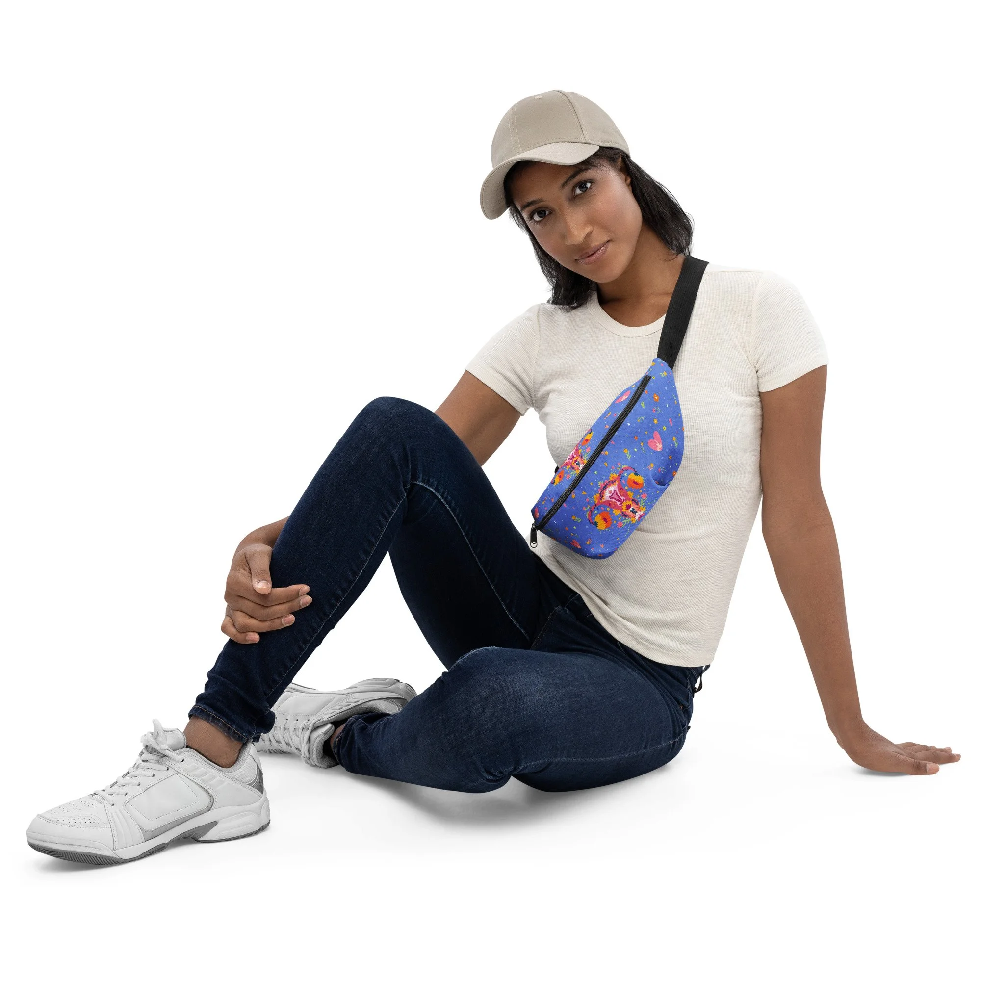 A woman wearing a beige baseball cap, white t-shirt, dark jeans, and white sneakers sitting on the floor with her legs crossed, holding a small colorful floral-patterned purse. Up Yours Menopause!' merchandise.