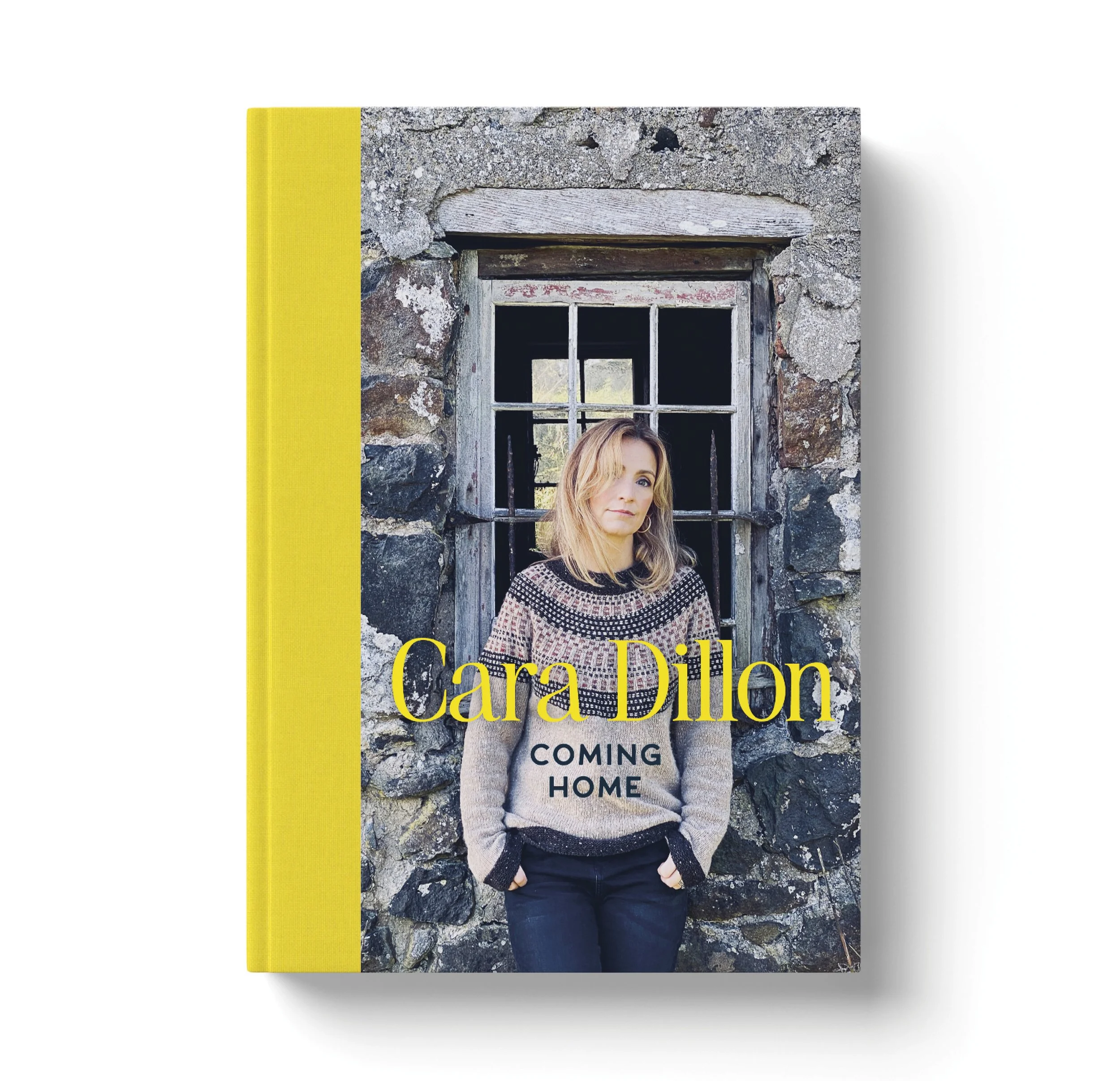 Book cover titled 'Coming Home' by Cara Dillon, featuring a woman with blonde hair wearing a patterned sweater, standing in front of a stone building with a window.