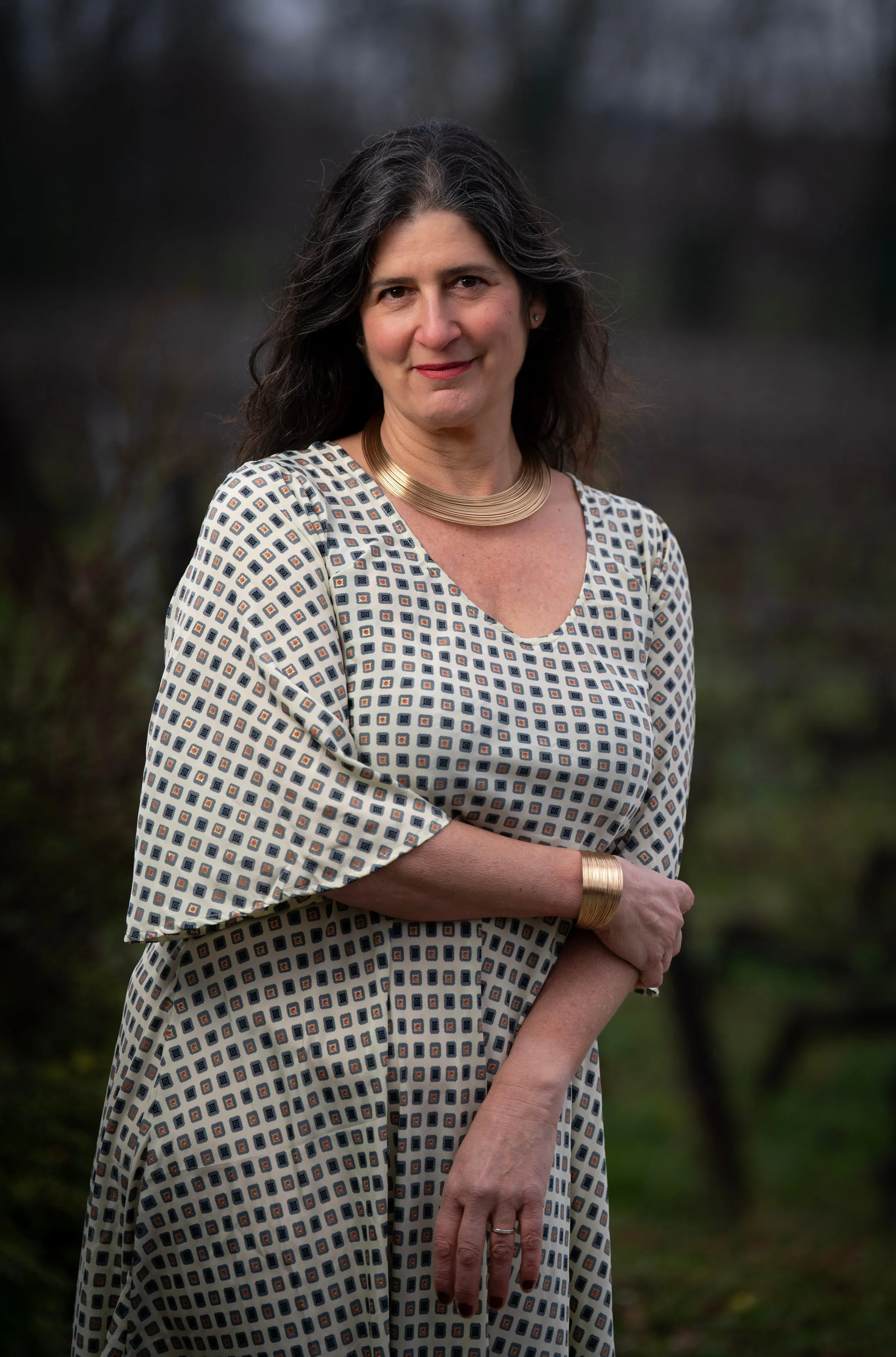A middle-aged woman with dark wavy hair, wearing a patterned dress with a v-neck, a gold necklace, a gold bracelet, and a ring, standing outdoors with a blurred nature background.