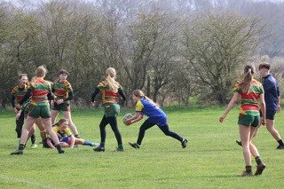 Women playing rugby on a grassy field, with some players tackling and others preparing to pass or receive the ball, overcast sky, trees in the background.