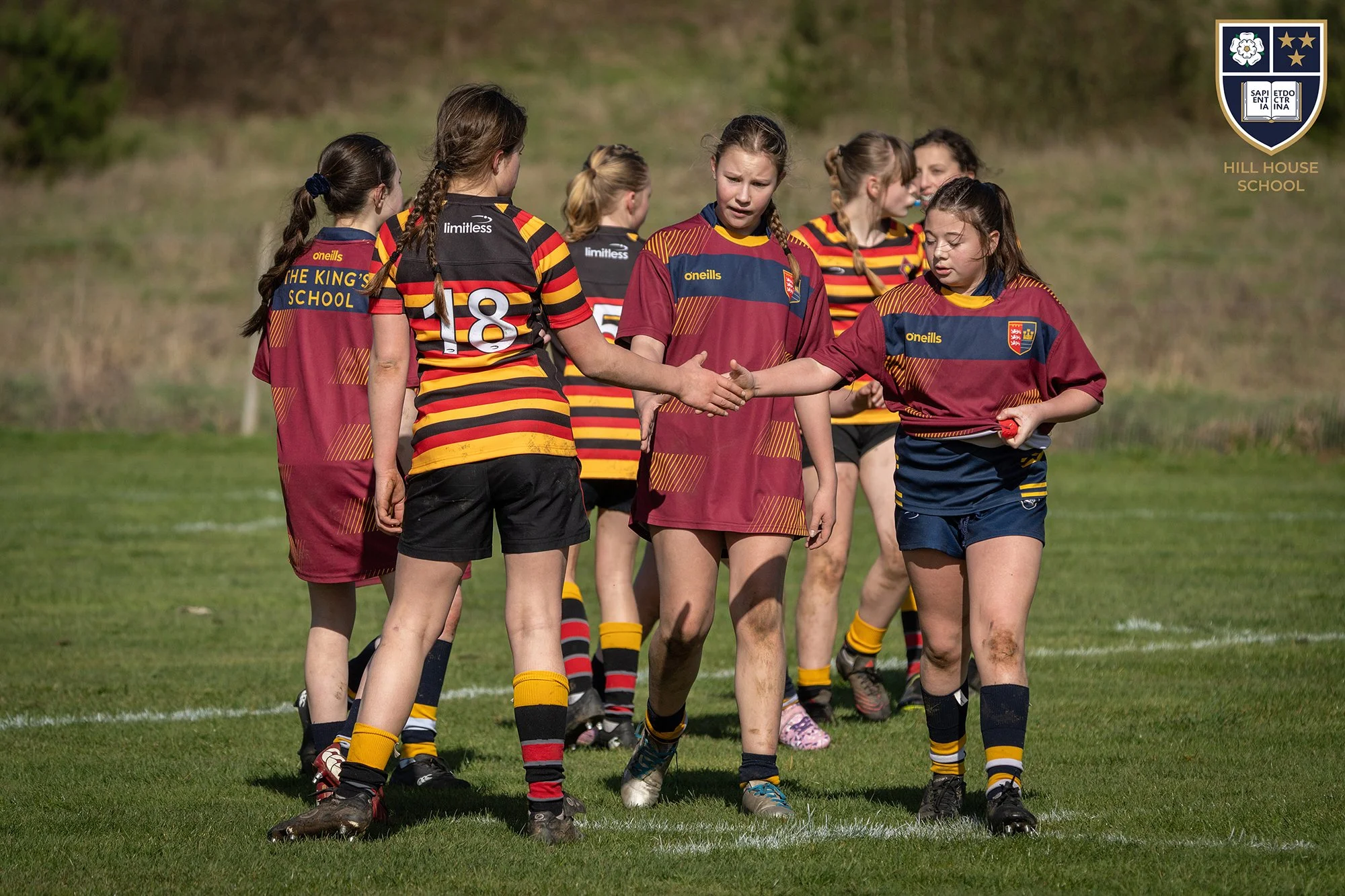 Young girls on a rugby field, shaking hands before a match, wearing maroon and black uniforms with yellow accents, with a grassy field and trees in the background.