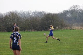 A girl running to catch a rugby ball during a game on a grassy field with three other players watching.