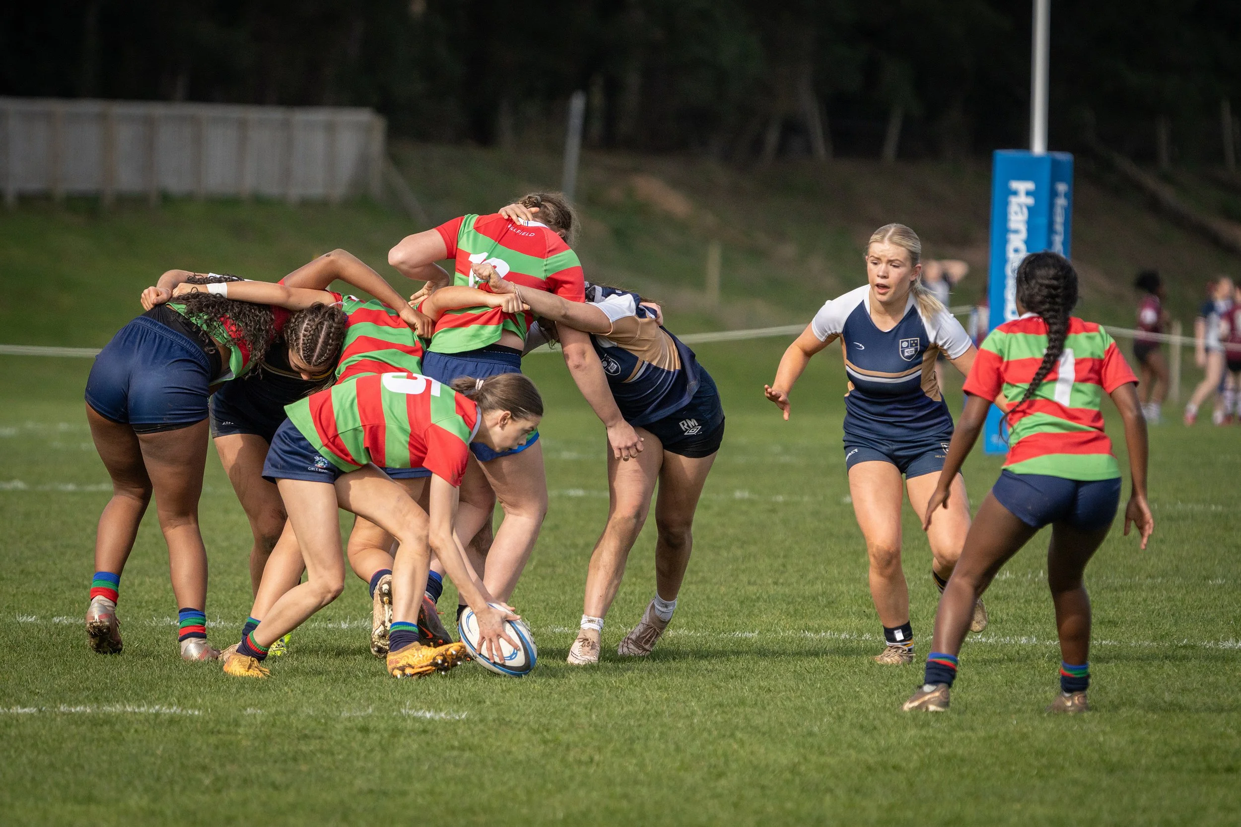 Women playing rugby on a field, with some players in blue uniforms and others in red and green striped jerseys, engaging in a scrum to gain control of the rugby ball.