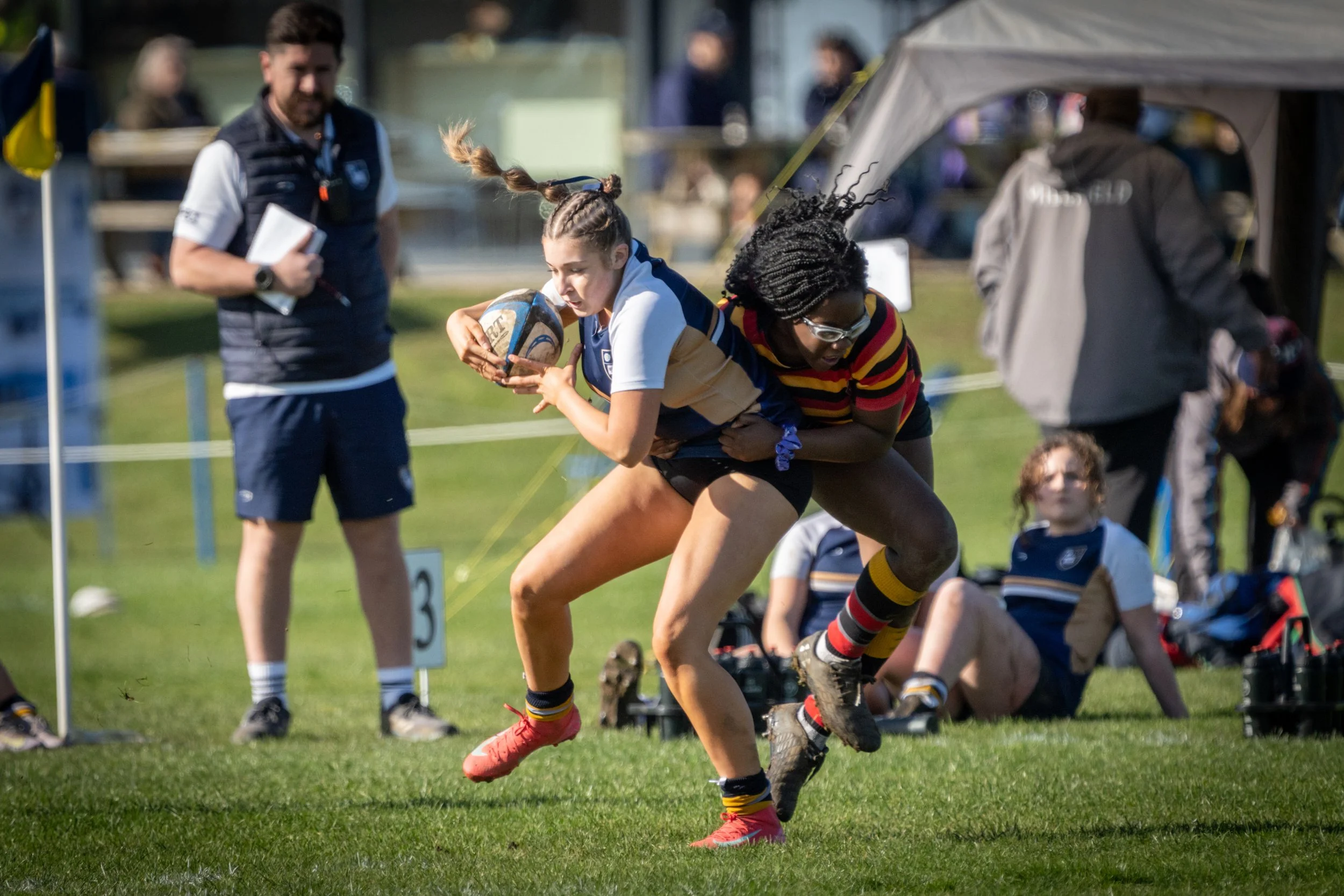 A rugby game with a female player in a white and blue jersey carrying the ball, tackled by another female player in a striped shirt, during a game on a grassy field with spectators and officials in the background.