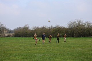 Four people playing rugby on a grassy field with trees and overcast sky in the background.