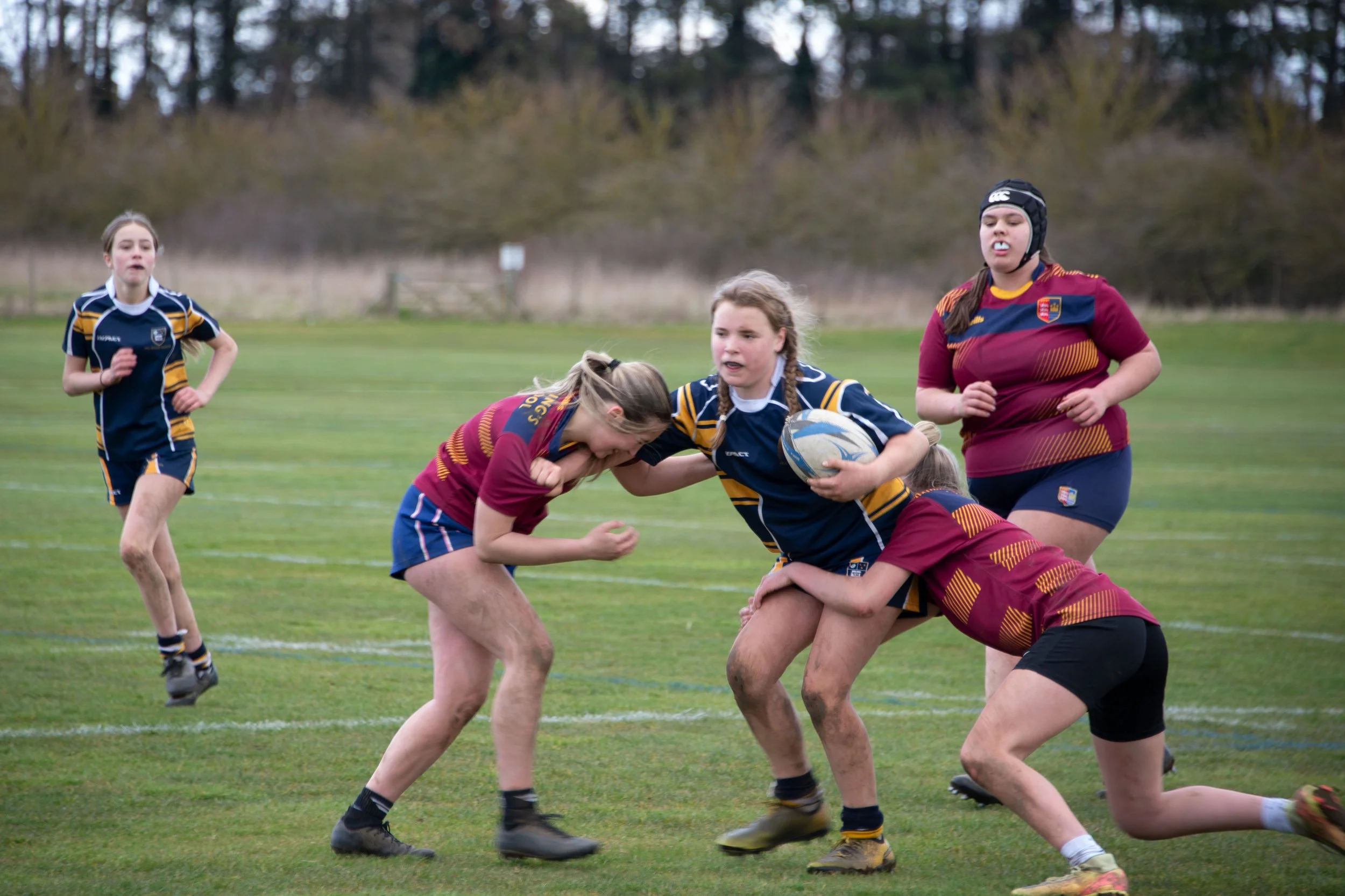 Young girls playing rugby on a grassy field, engaging in a tackle during a game, with trees in the background.