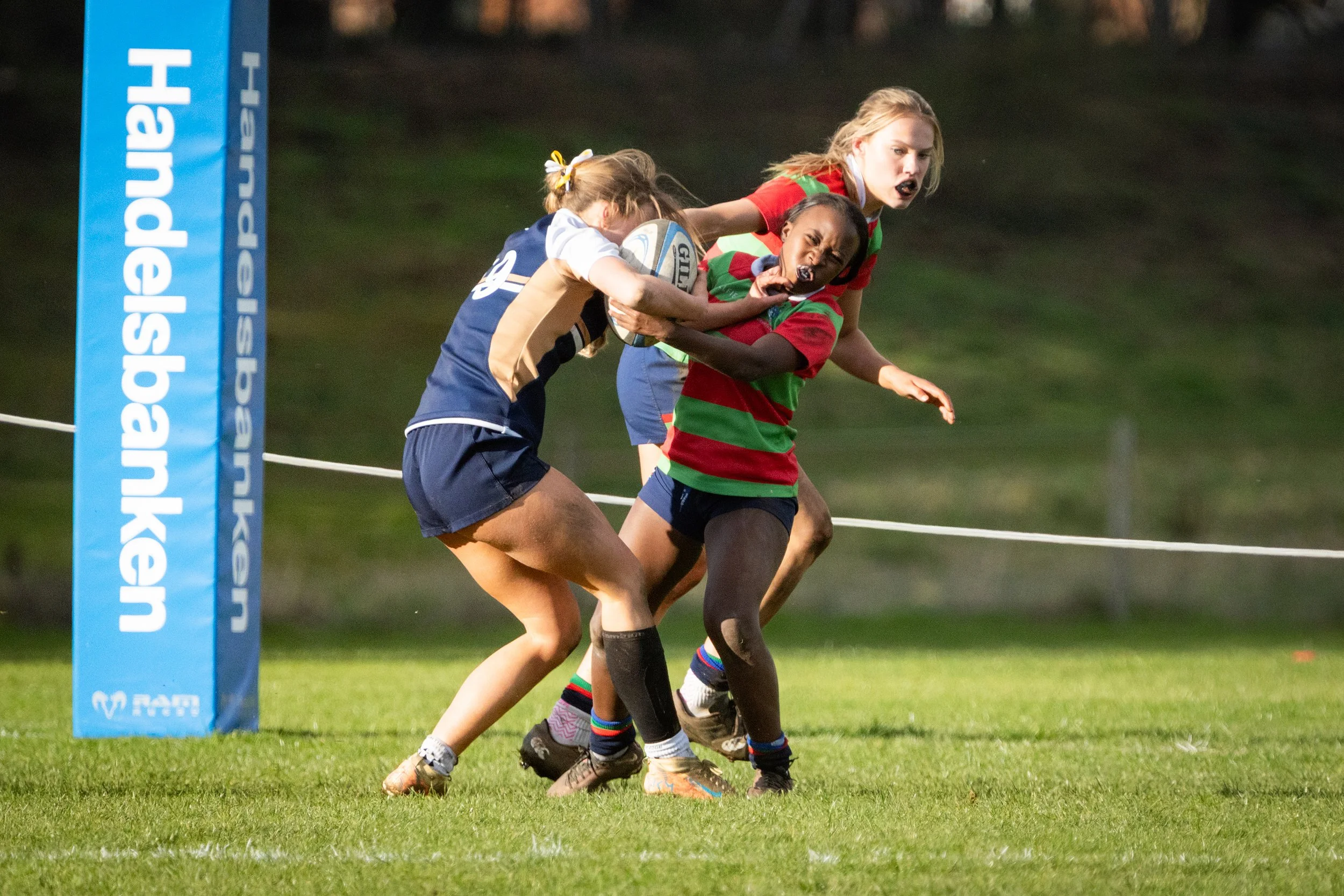 Young girls playing rugby on a field near a rugby goalpost, one girl holding a rugby ball while being tackled by another girl, with a logo on the goalpost reading 'kansaskalder.com'.