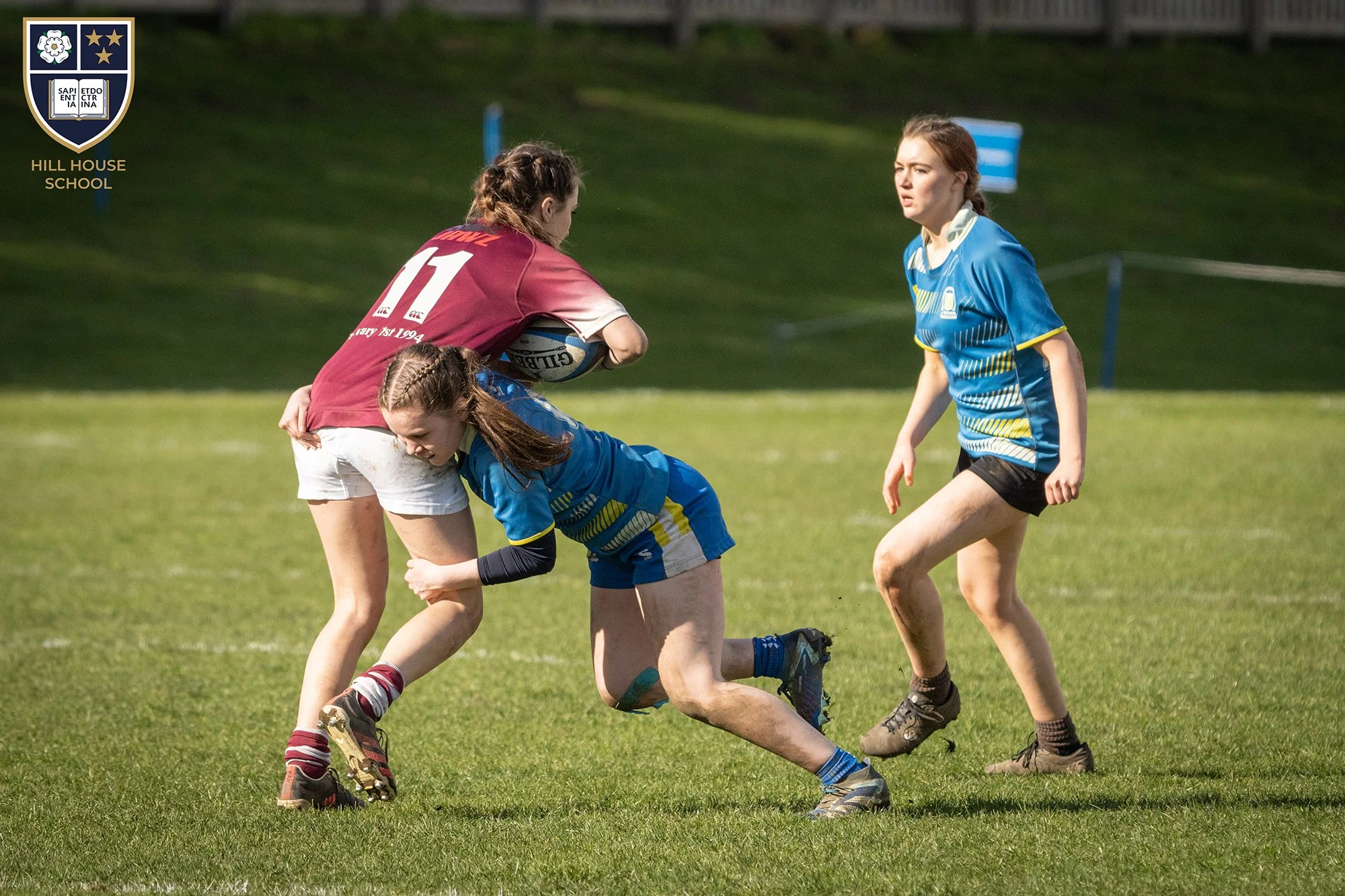 Young girls playing rugby on a grassy field, one holding a rugby ball, other girls in blue uniforms preparing to engage.