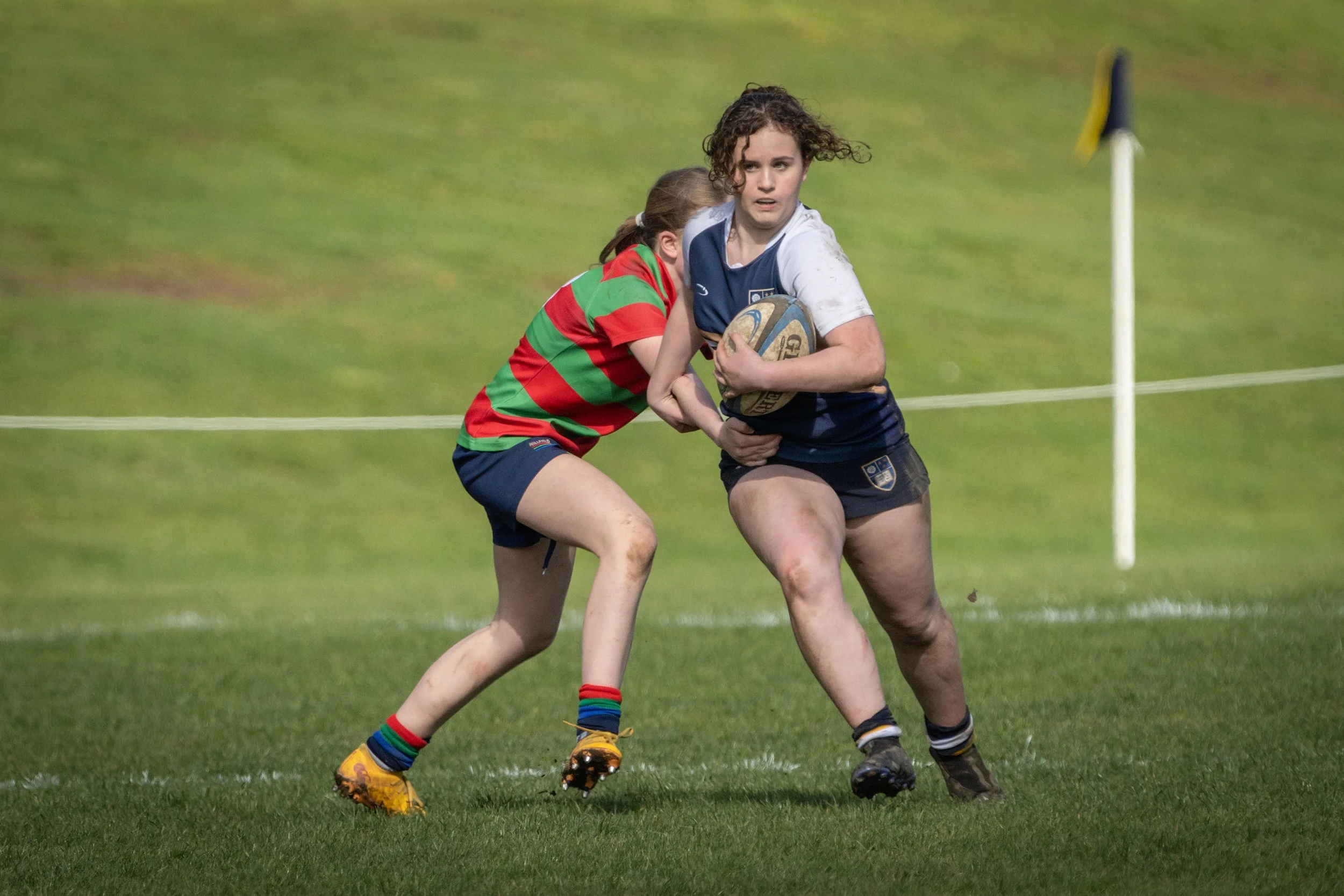 Two women playing rugby on a grassy field, one in a blue and white uniform holding a rugby ball, and the other in a red and green striped shirt trying to tackle her.