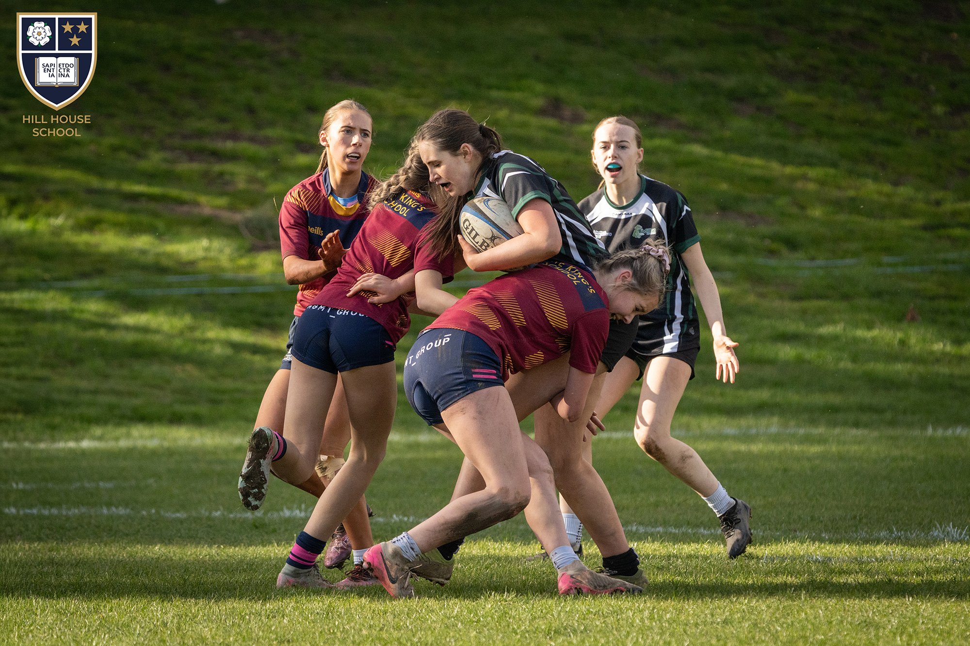 Girls playing rugby on a grassy field during daytime, wearing sports uniforms and one holding a rugby ball.