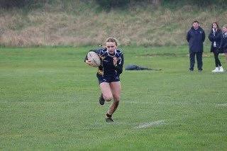 A person running on a grassy field while holding a rugby ball, with two spectators in the background.