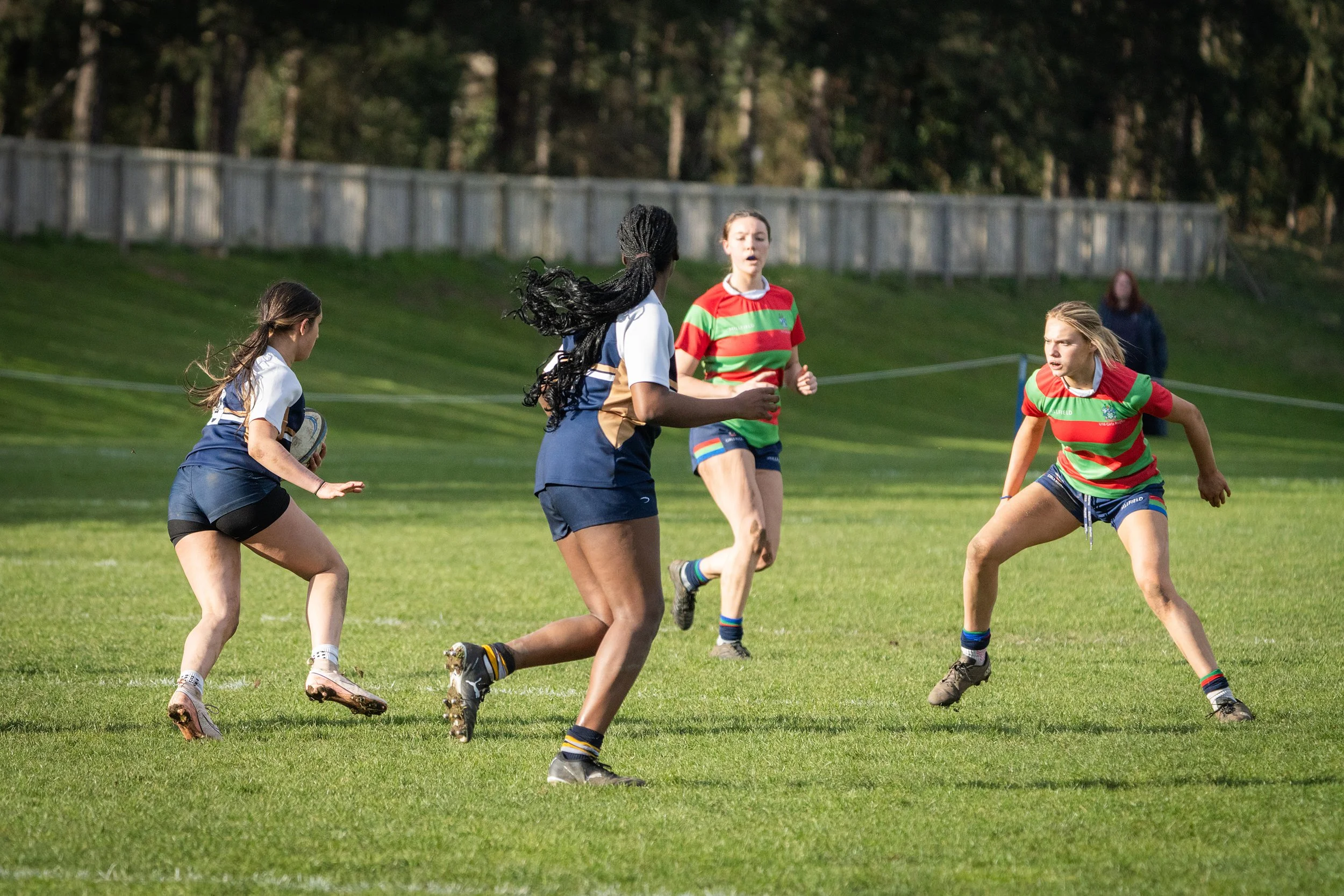 Women playing rugby on a grassy field with a wooden fence and trees in the background.