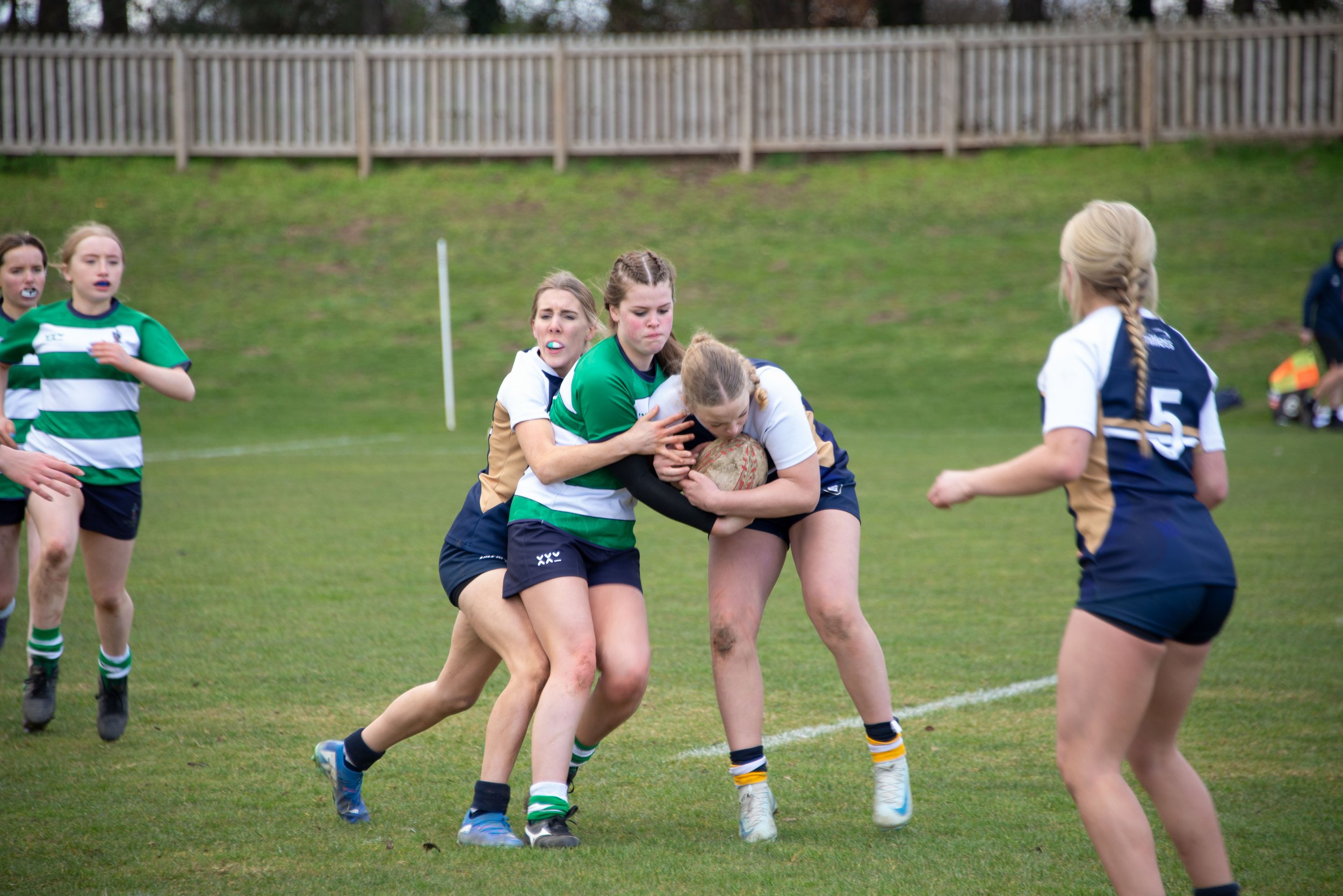Women playing rugby on a grassy field, with one woman tackling another with the ball, while others observe or participate in the game.