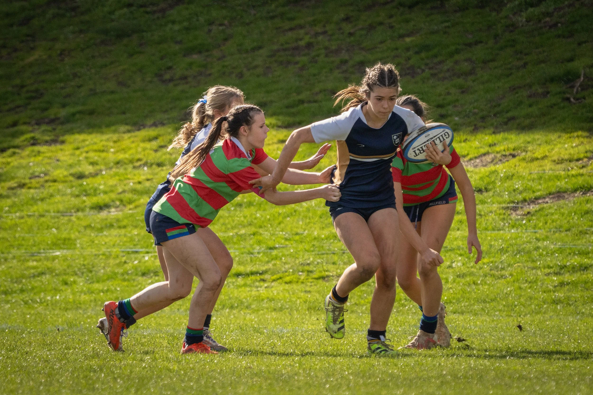 Young women playing rugby on grassy field, one carrying ball while others attempt to stop her.