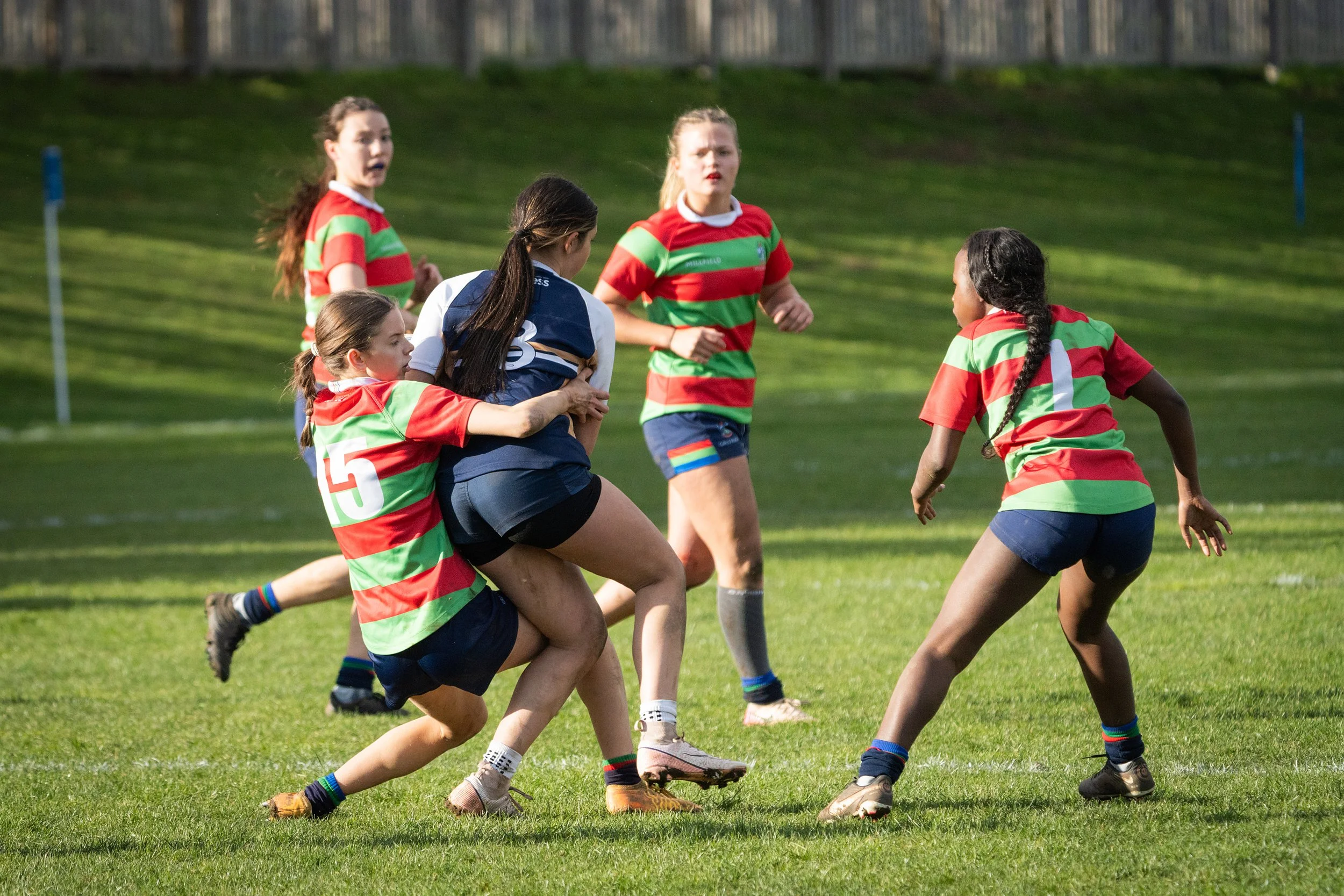 Girls playing rugby in a grassy field, some are tackled while others prepare for the next move, wearing colorful jerseys.