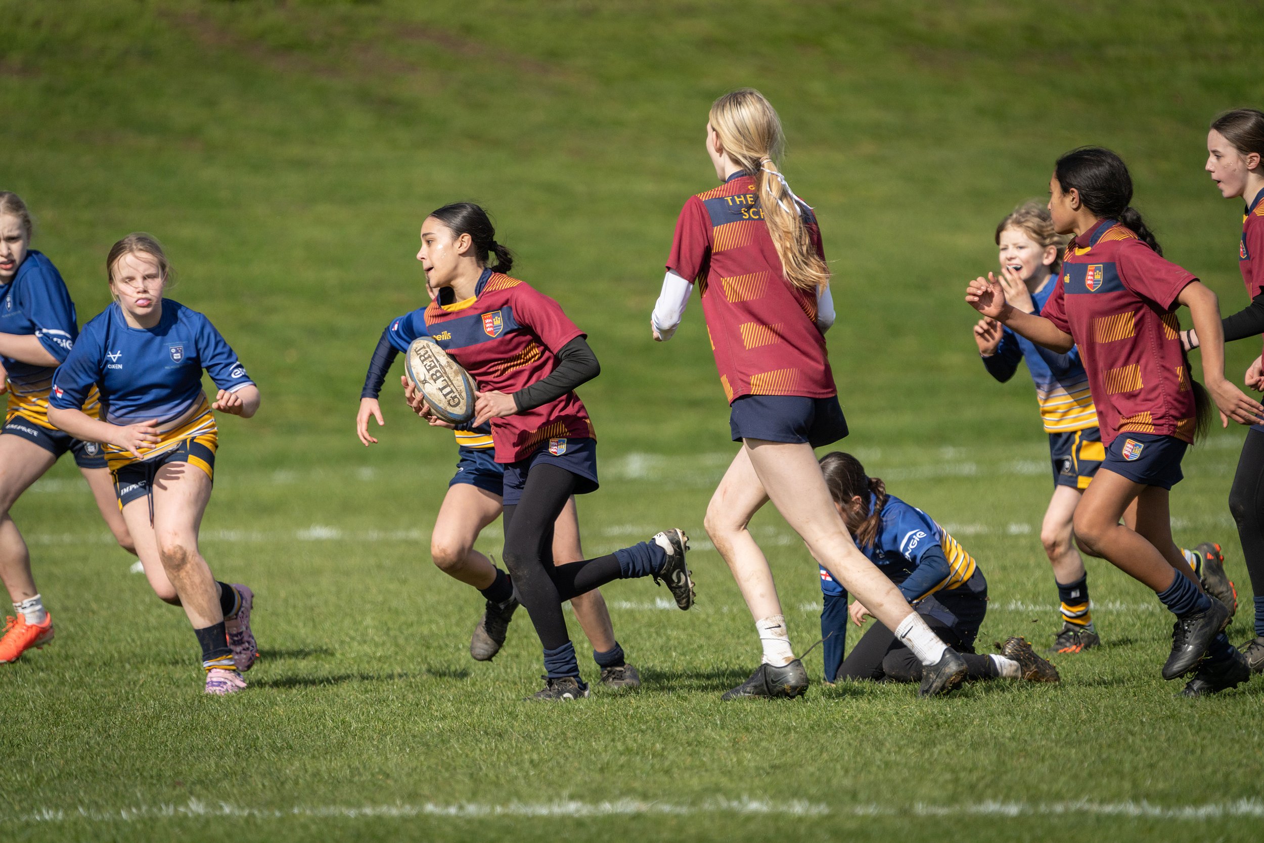 Young girls playing rugby on a grassy field, wearing colorful sports uniforms.