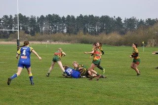 Women playing rugby on a grassy field with trees in the background; one player is tackled to the ground while others reach out to help or defend.