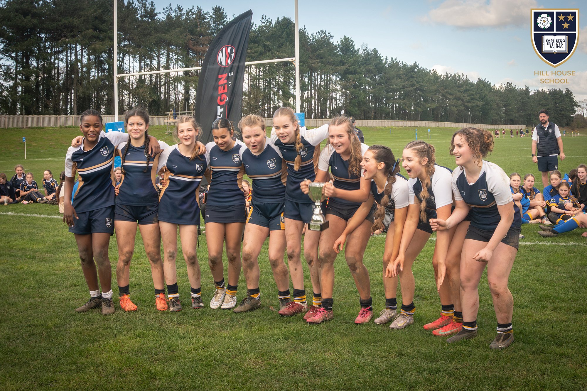 A group of young female rugby players in navy and gold uniforms celebrating on a grassy field after a game, with some muddy legs, holding a trophy, with a rugby goalpost, a large black flag, and other players in the background.