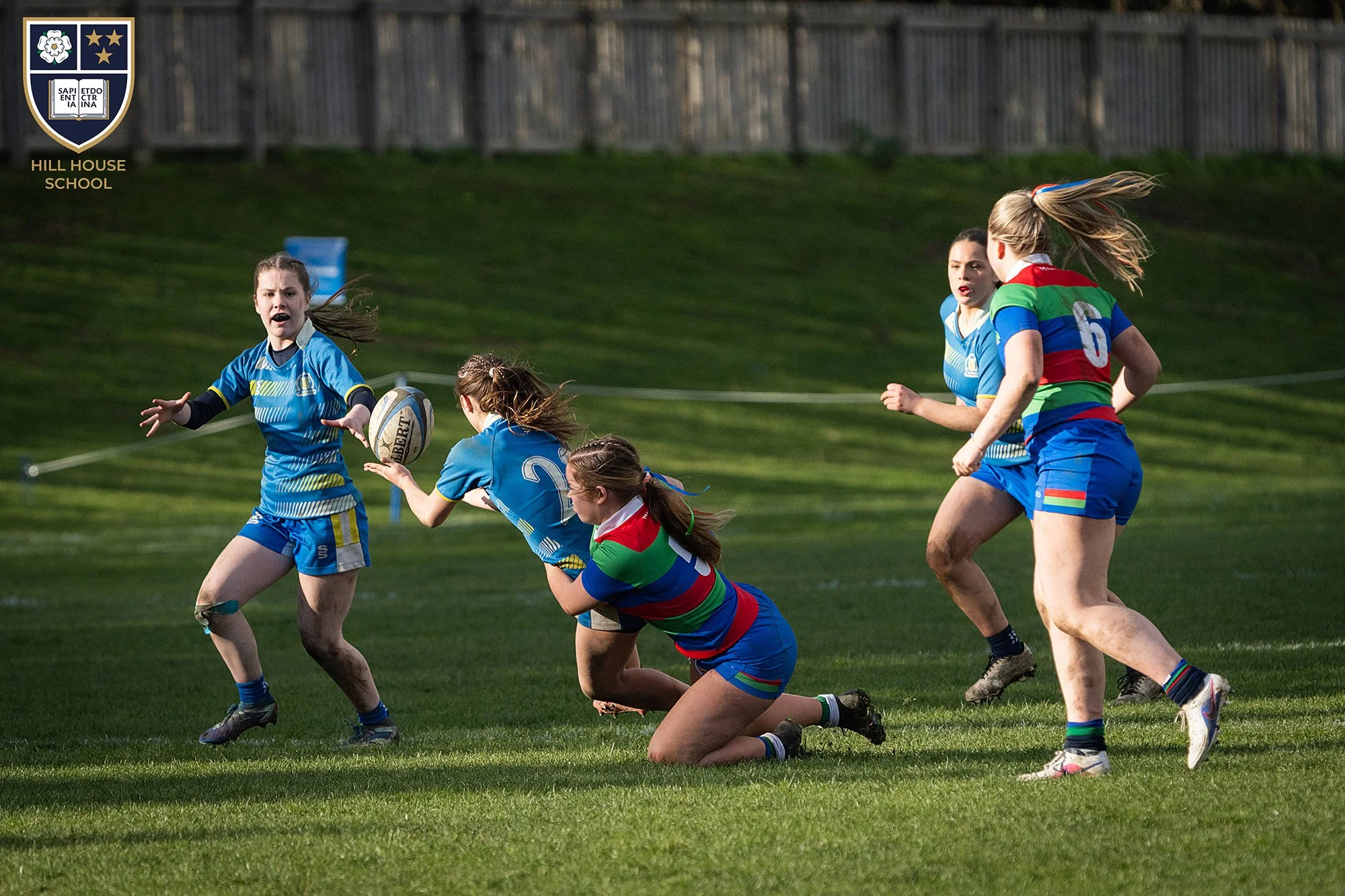 Girls playing rugby on a grassy field, some kneeling and others running, wearing colorful sports uniforms, with a fence in the background and the Hill House School logo in the top left corner.